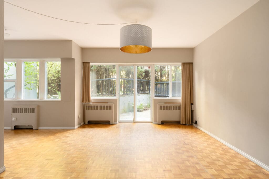 Empty room with light parquet floors, beige walls, ceiling light, large windows, and glass door to patio with trees in Vancouver.