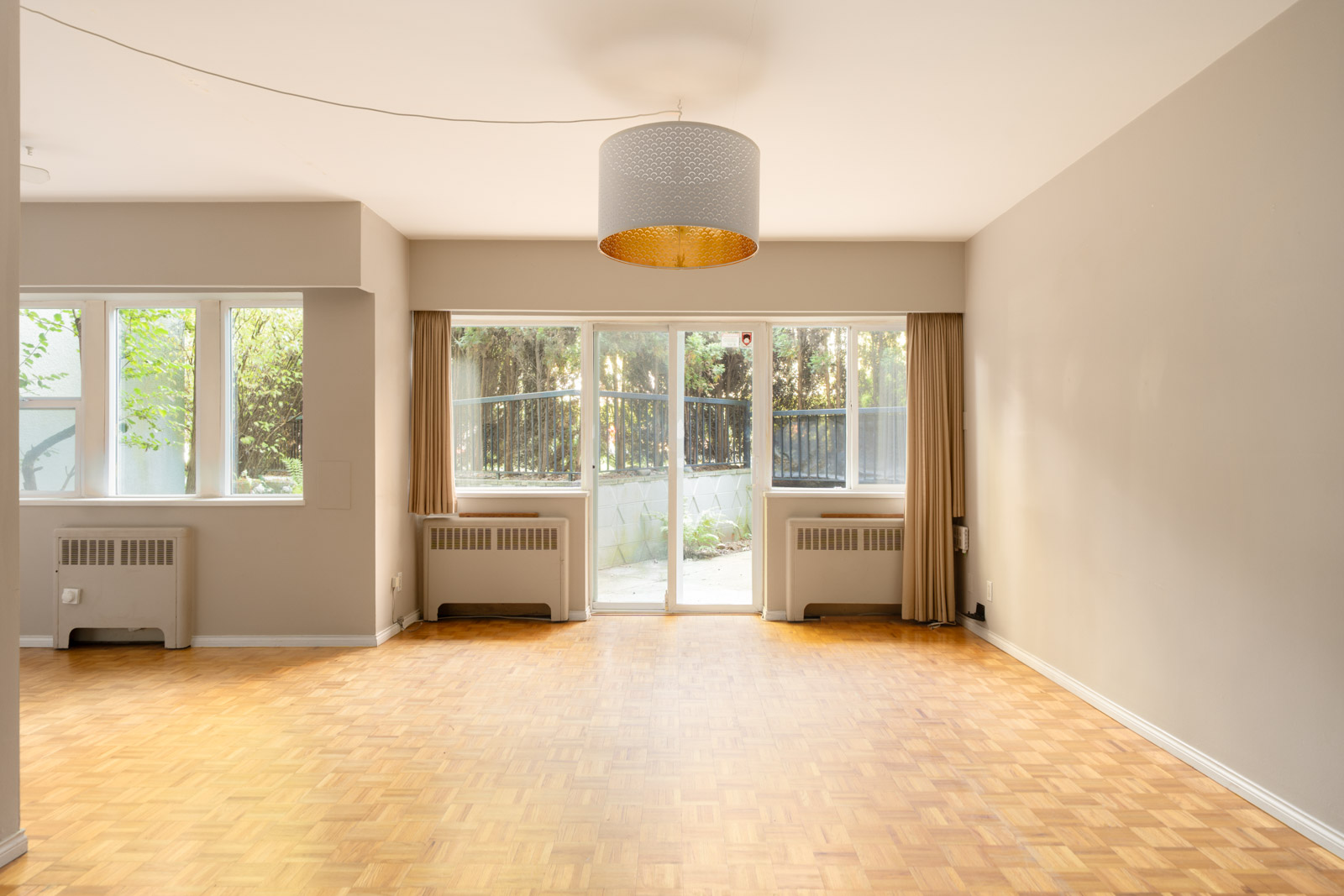 Empty room with light parquet floors, beige walls, ceiling light, large windows, and glass door to patio with trees in Vancouver.