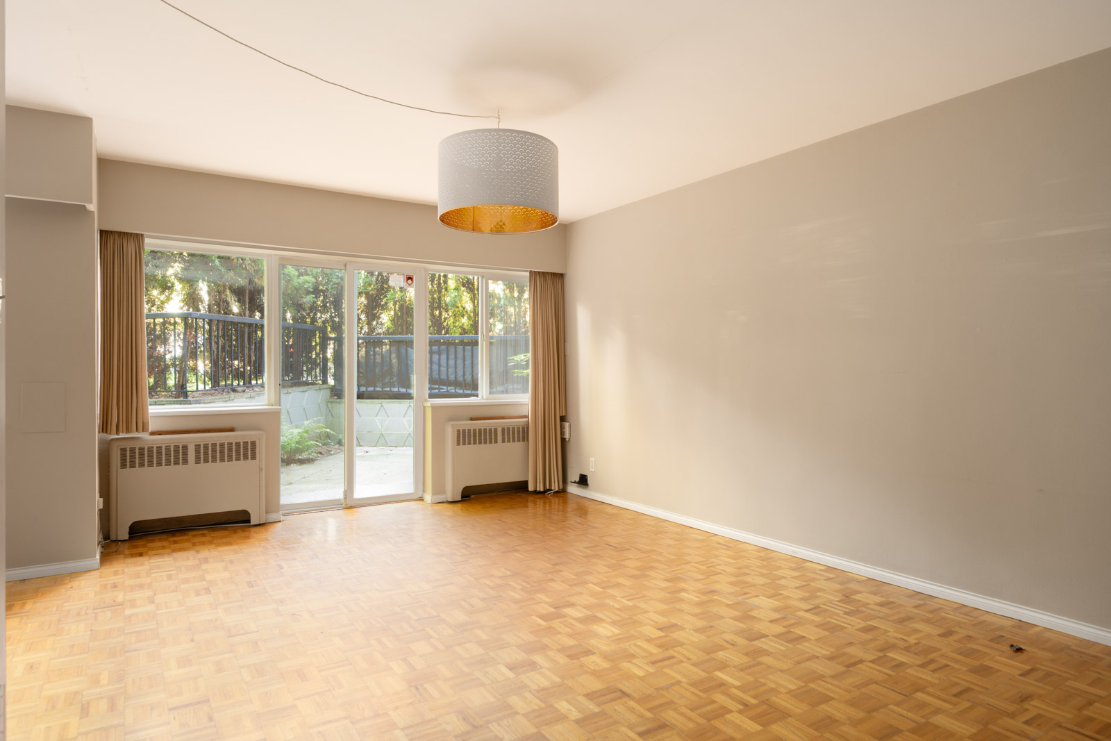 Empty living room at 1970 W 41st Ave, Vancouver, with parquet floors, beige walls, big windowed doors, two radiators, and ceiling light.
