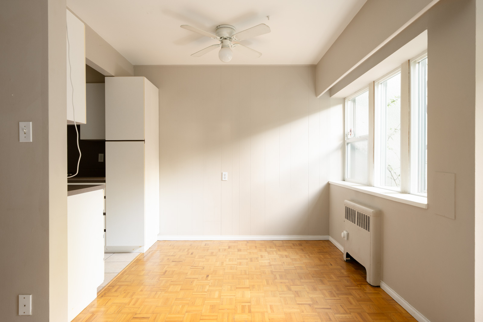 Empty room with parquet floors, large window, ceiling fan, radiator, and partial kitchen view in a Vancouver residence.