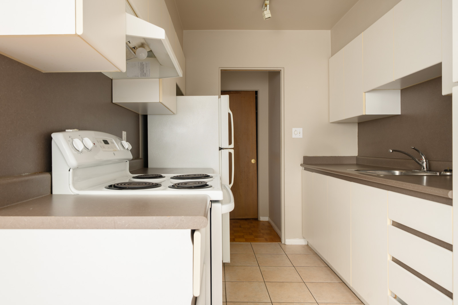 Narrow kitchen with white cabinets, brown counters, electric stove, sink, and refrigerator. Door at the end. Vancouver location.
