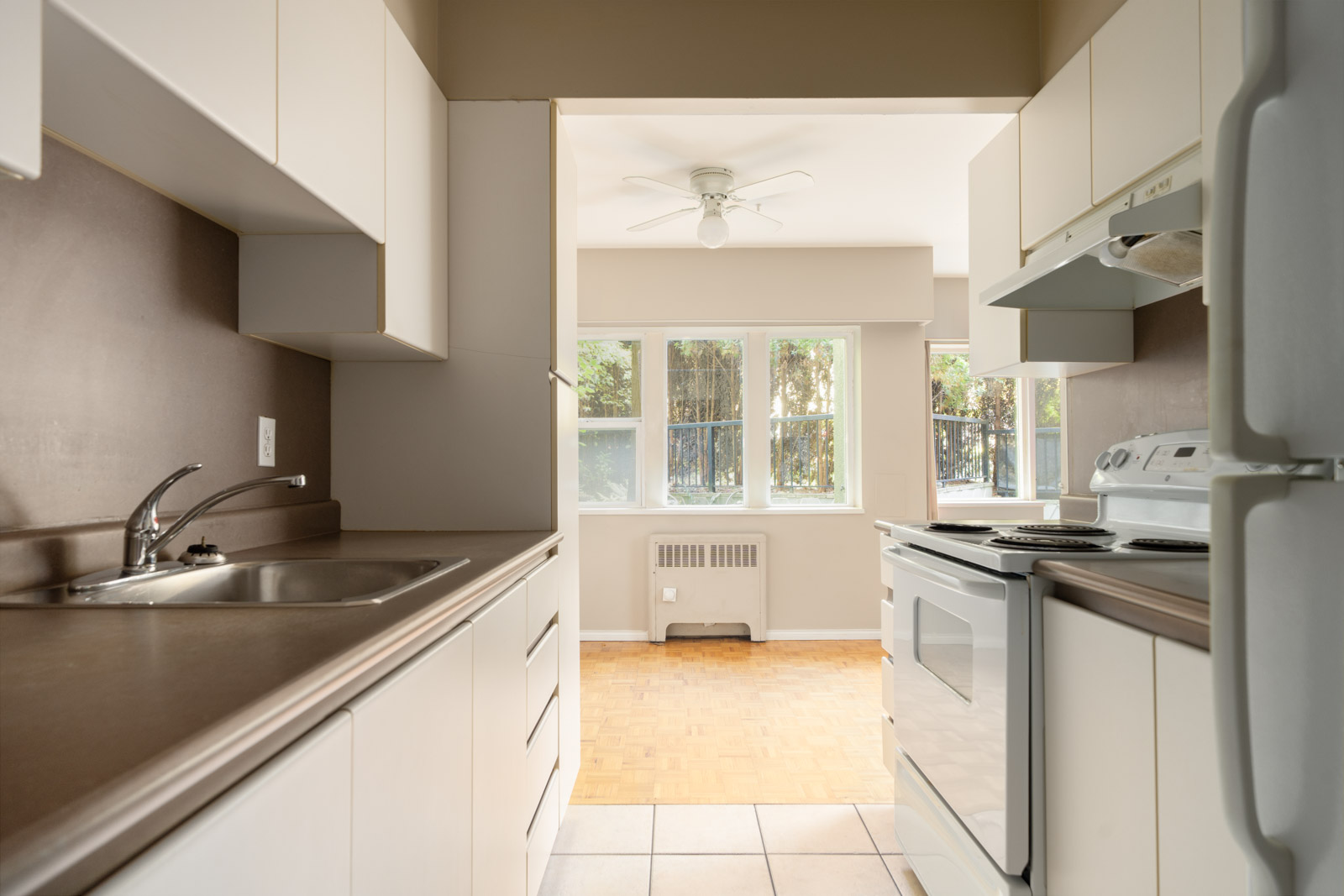 Narrow kitchen with white cabinets, electric stove, and sink, opening to a bright dining area with large windows and ceiling fan in Vancouver.
