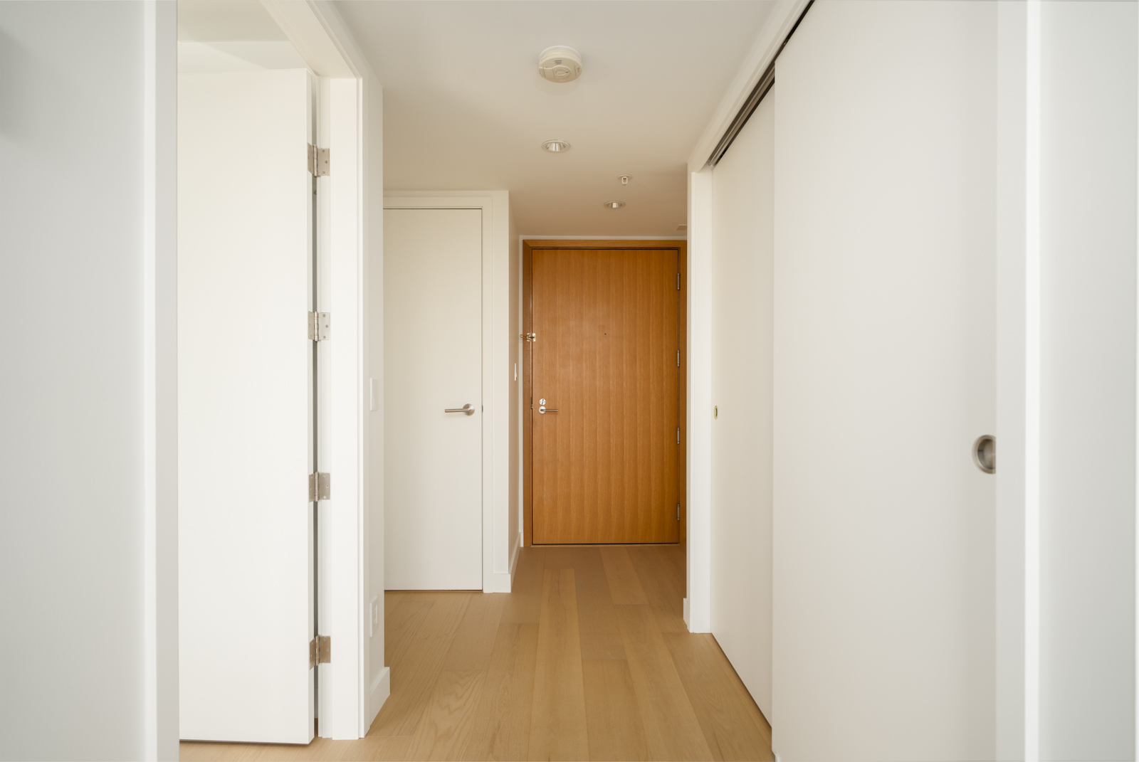 A bright, minimalist hallway at 1565 West 6th Ave, Vancouver, with light wood floors, white walls, and a wooden front door.