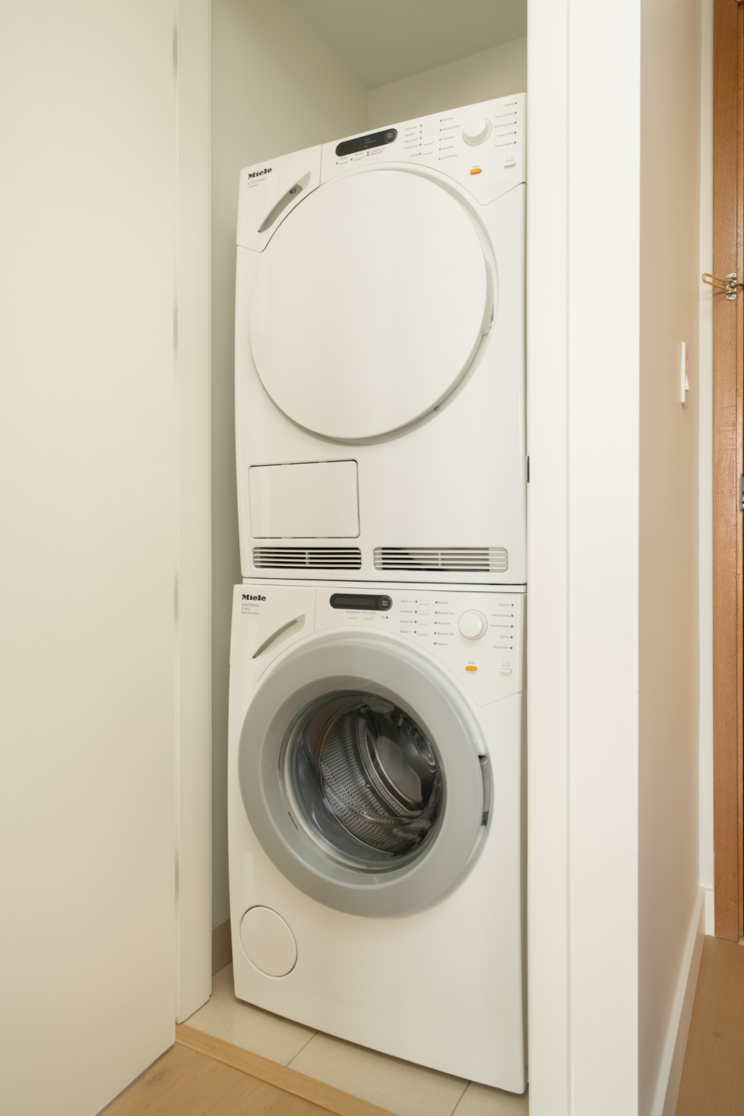 Stacked front-loading washer and dryer in a narrow white-walled laundry closet with wood floor, 11th Floor, Vancouver.