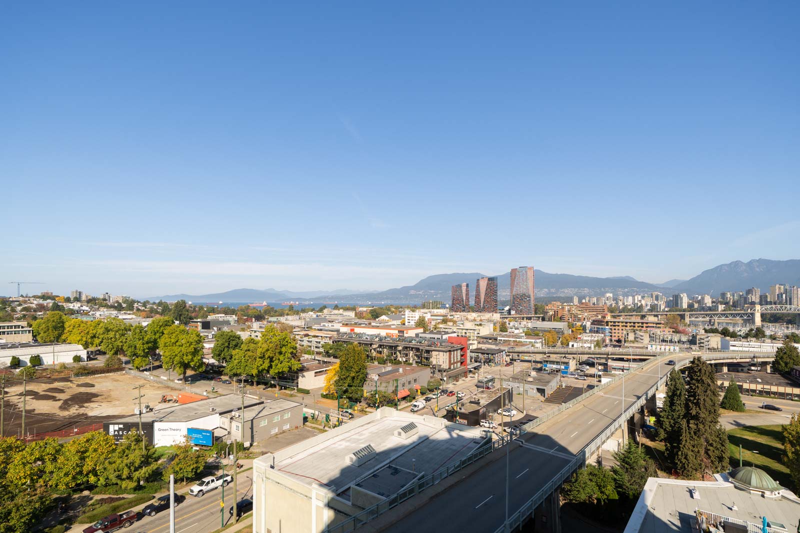 Aerial view of Vancouver’s streets, buildings, and trees beneath a clear sky, with mountains behind 1565 West 6th Avenue.