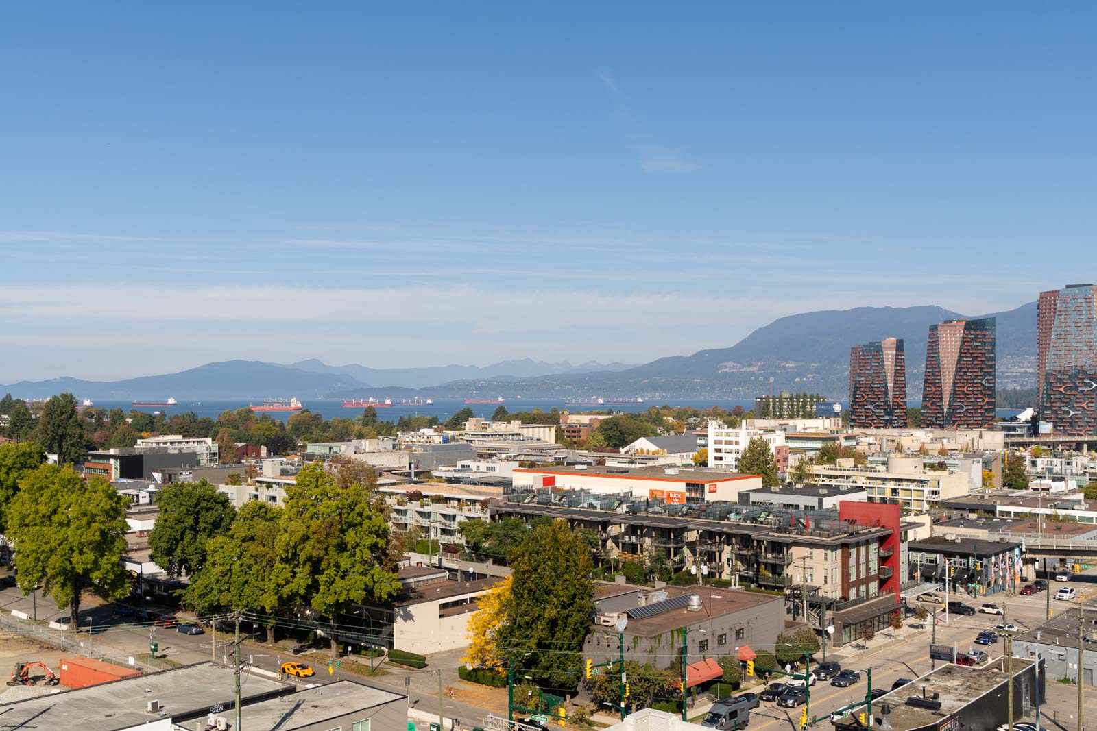 View from 11th floor at 1565 West 6th Ave, Vancouver: low buildings, trees, mountains, and ships on blue water under clear sky.