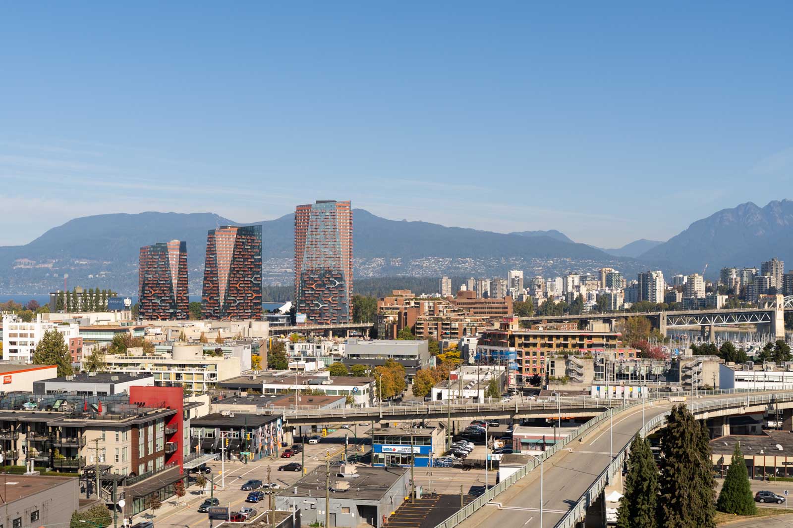 Vancouver cityscape with high-rises, low buildings, roads, and distant mountains under clear sky, seen from 1565 W 6th Ave, 11th floor.