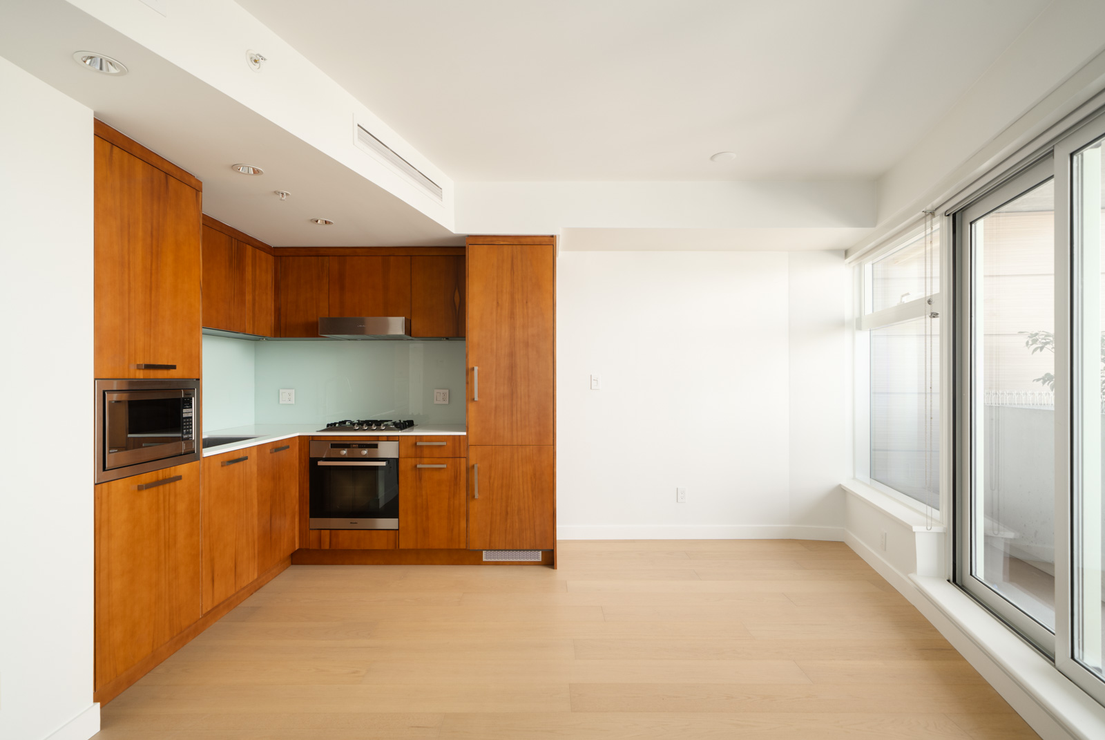 Modern kitchen with wood cabinets, built-in appliances, light wood floors, large windows, white walls; 11th floor in Vancouver.