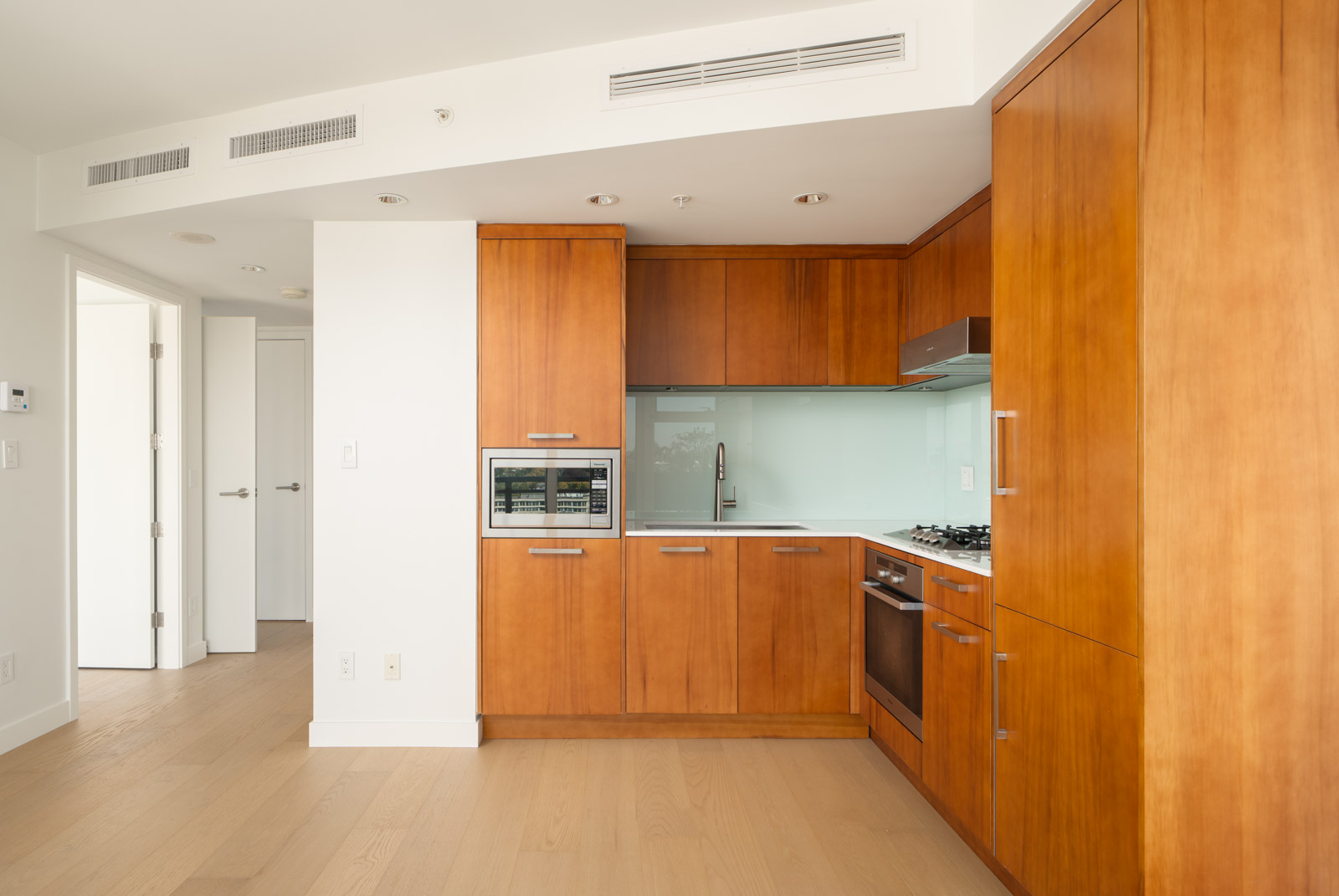 Modern kitchen with wooden cabinets, built-in oven, gas stove, microwave, glass backsplash, and two open doors. Vancouver.