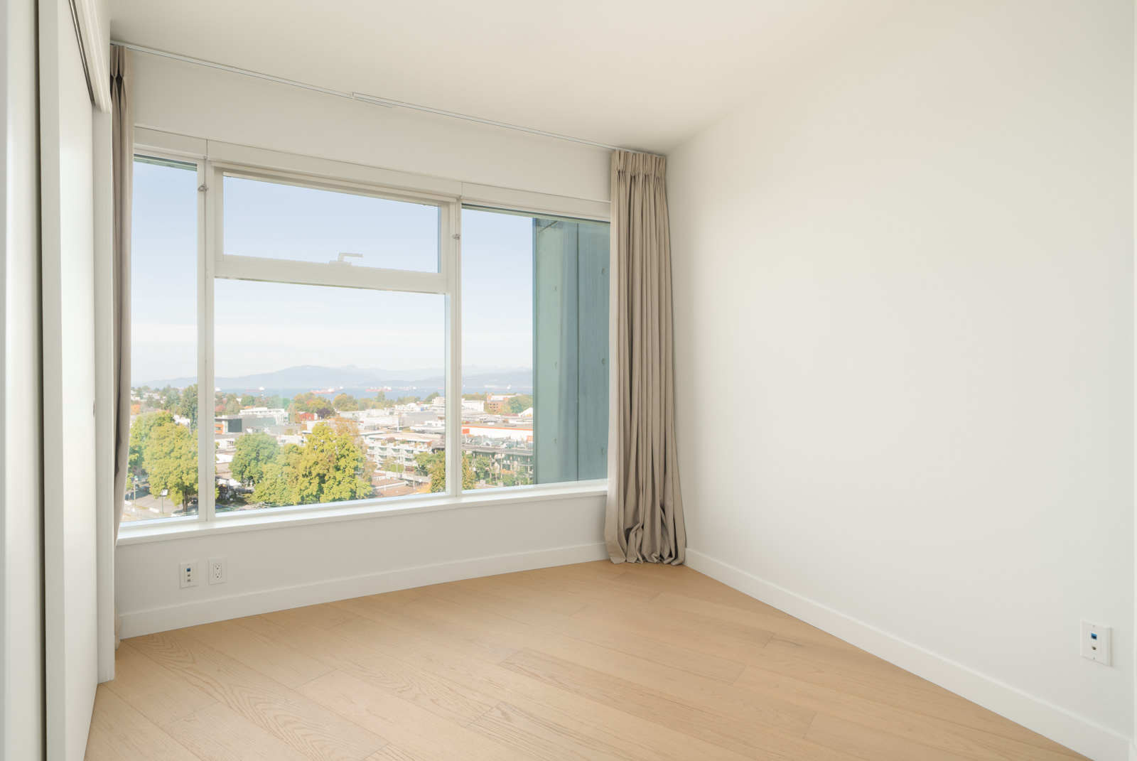 Bright, unfurnished room with light wood floors, white walls, beige curtains, and a large window overlooking Vancouver and mountains.