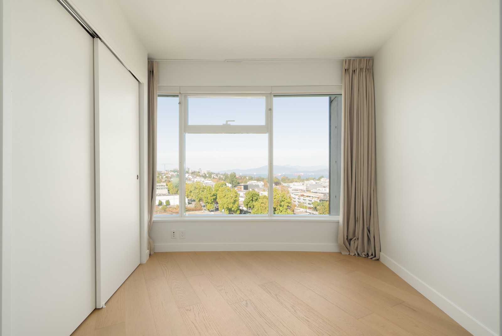 Bright empty room with light wood flooring, large window, white closet door; city and trees visible from 11th floor in Vancouver.