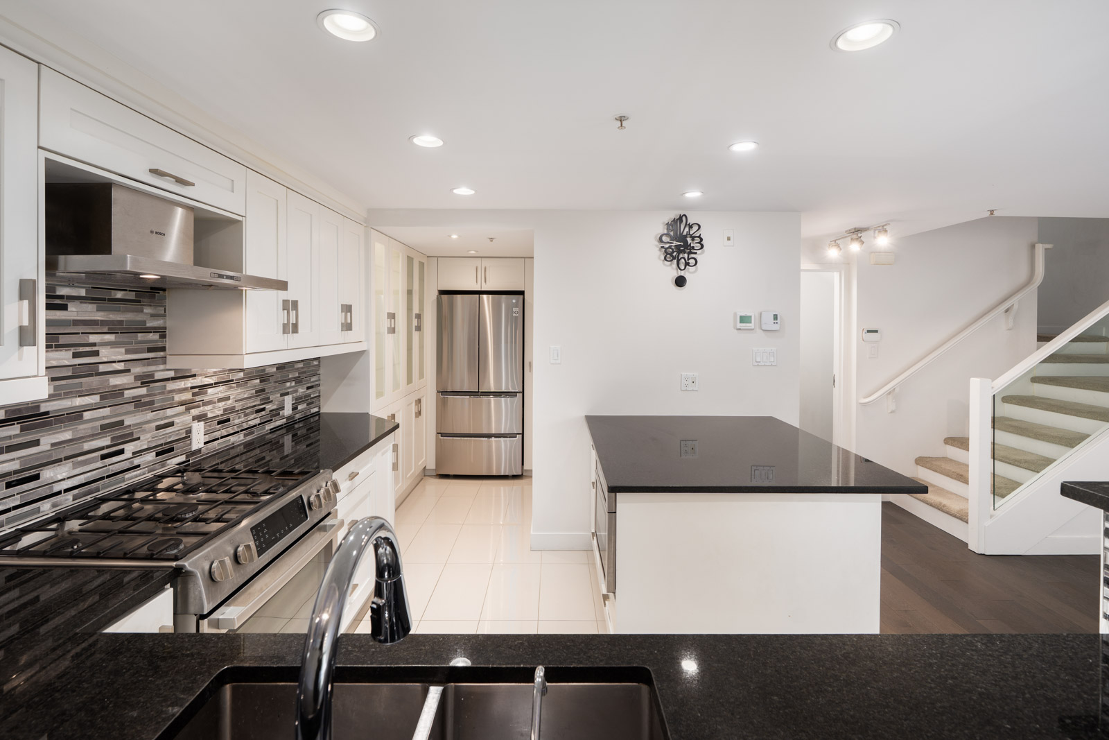 Modern kitchen with stainless appliances, white cabinets, black countertops, island, and staircase in a Vancouver townhouse.