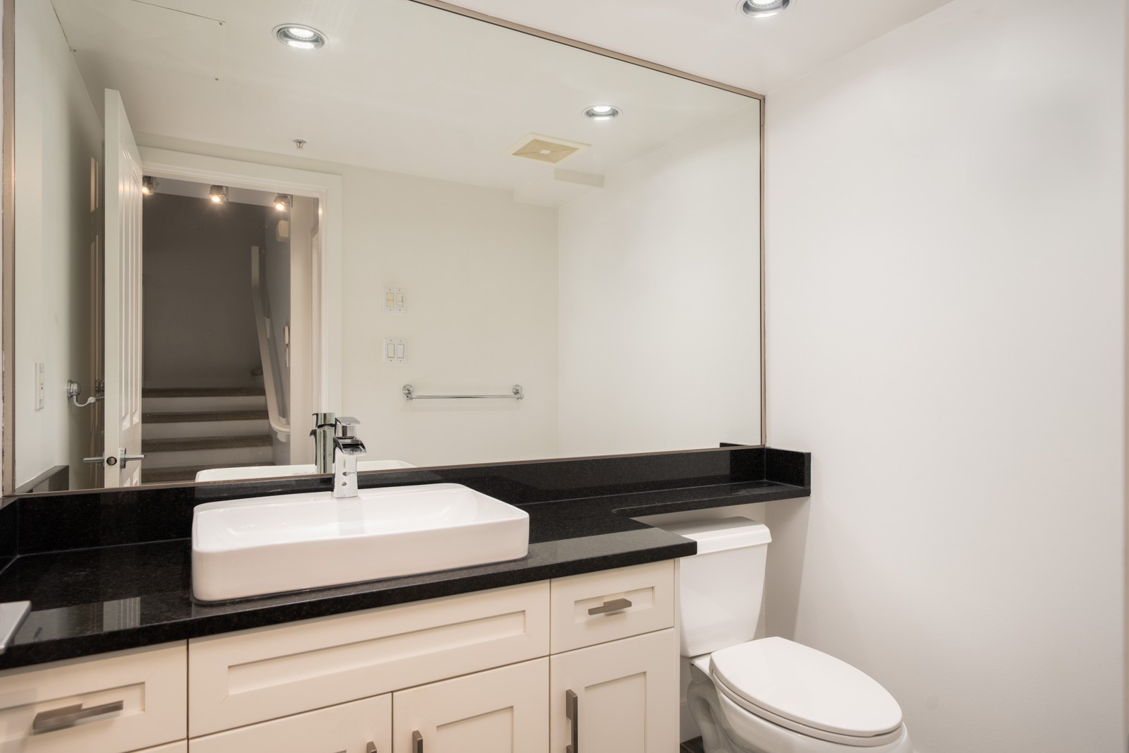Modern bathroom with white walls, black countertop, rectangular sink, toilet, and large mirror reflecting a door and staircase in Vancouver.