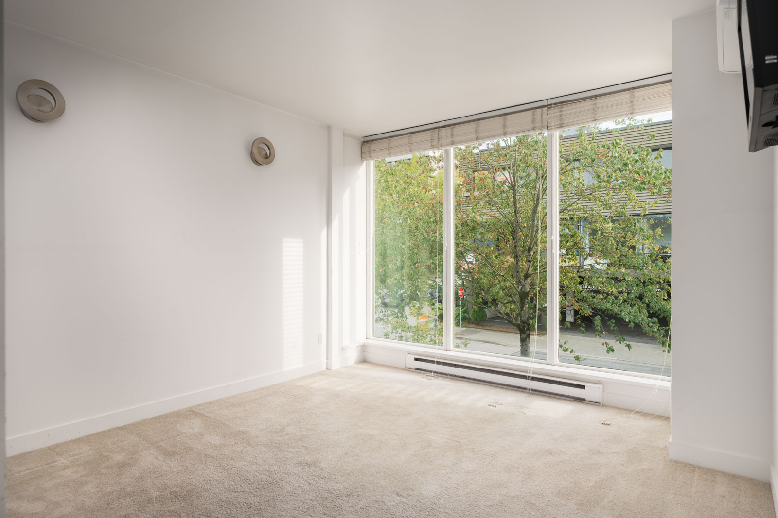 Empty room with beige carpet, white walls, two wall lights, and tall windows overlooking trees in Vancouver at 1428 West 6th Ave.
