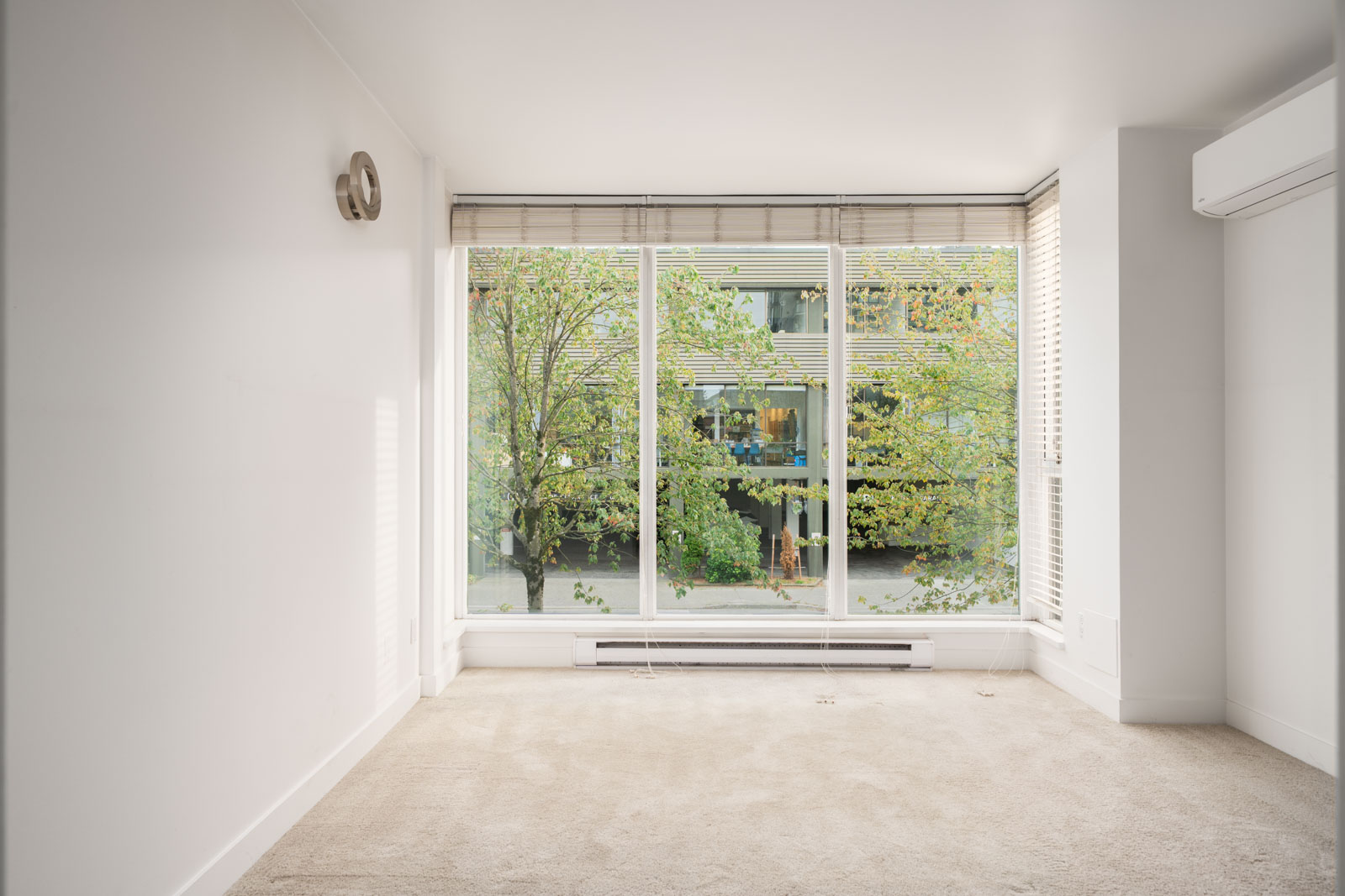 Empty room with beige carpet, white walls, floor-to-ceiling windows, and views of trees and a building in Vancouver.