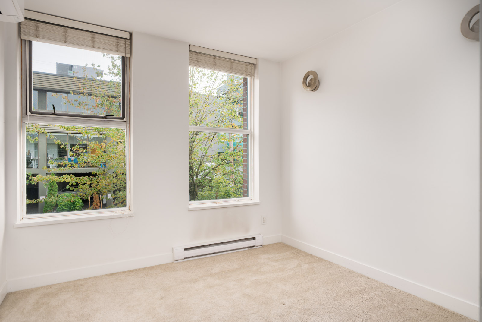 Empty room with beige carpet, white walls, two windows with blinds, wall heater, and view of trees and buildings in Vancouver.
