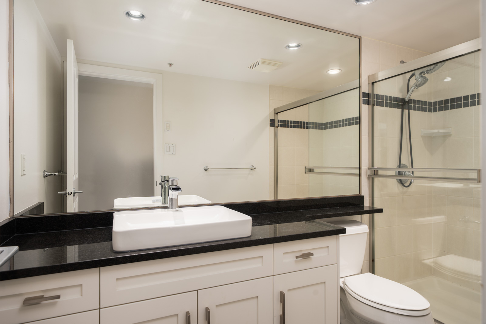 Modern bathroom in a Vancouver townhouse with a large mirror, white sink, black counter, white cabinets, toilet, and glass shower.