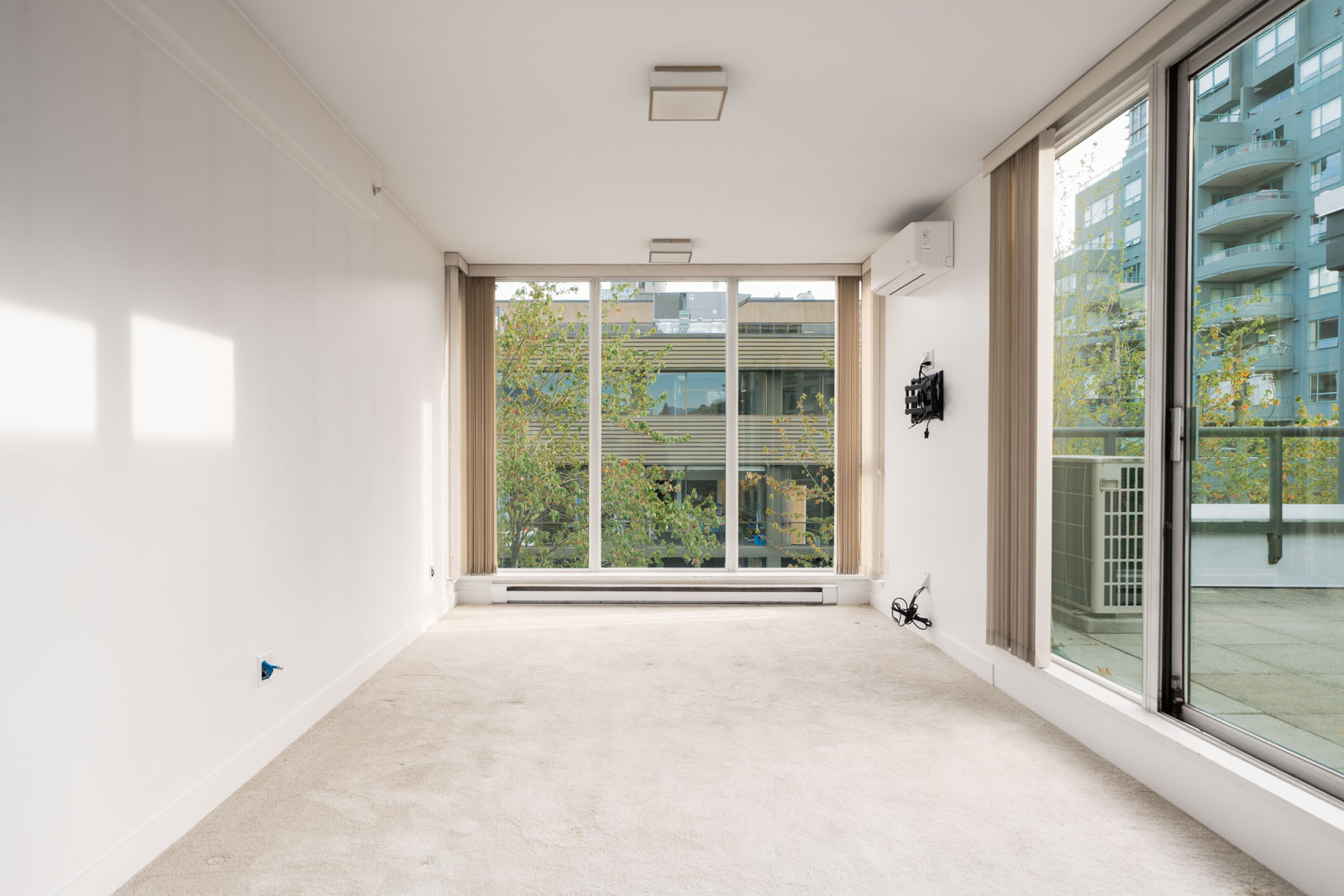 Bright, empty living room with beige carpet, large windows, sliding balcony door, and urban views in Vancouver on West 6th Avenue.
