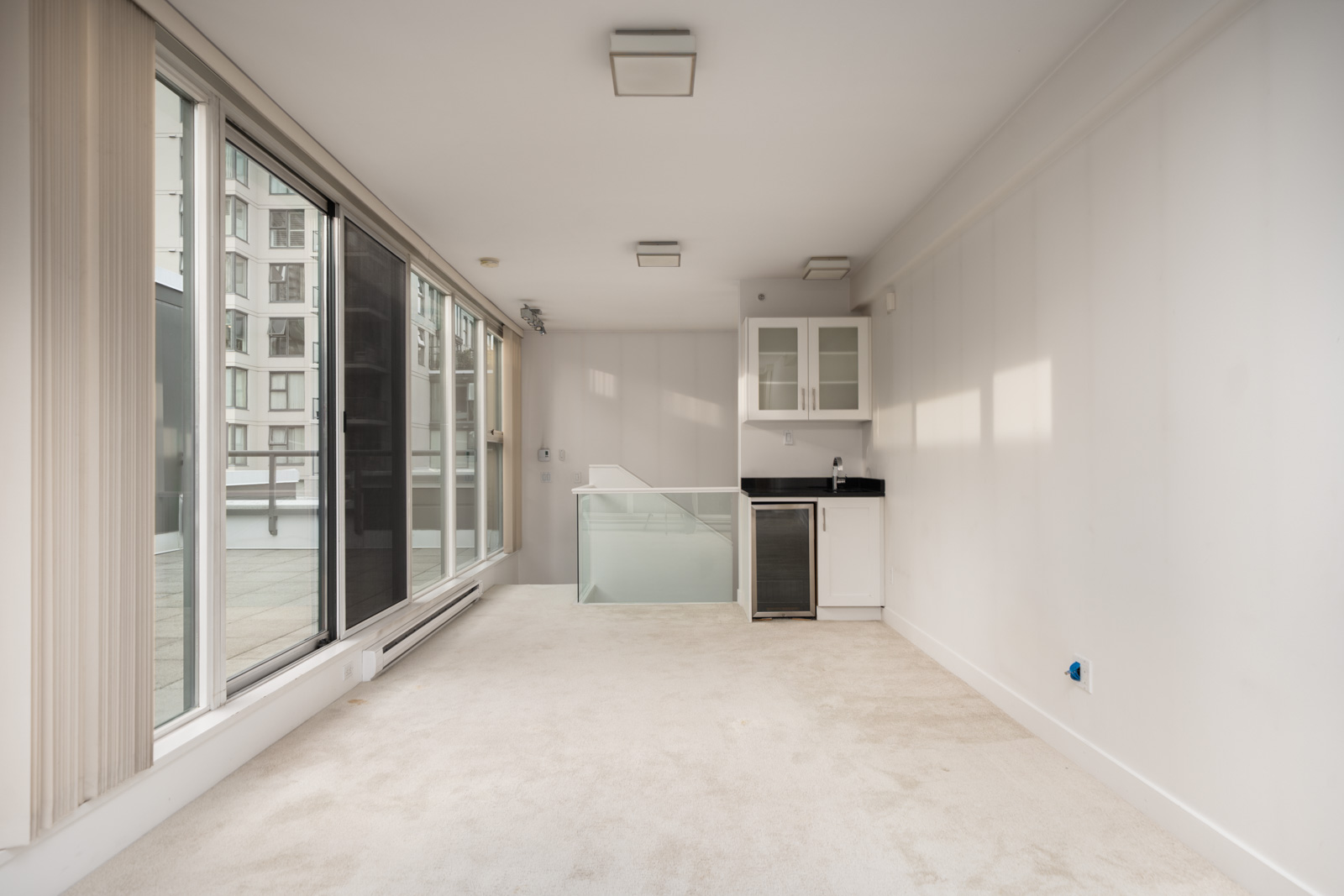 Unfurnished modern room in a Vancouver townhouse with large windows, white walls, beige carpet, kitchenette, and patio access.