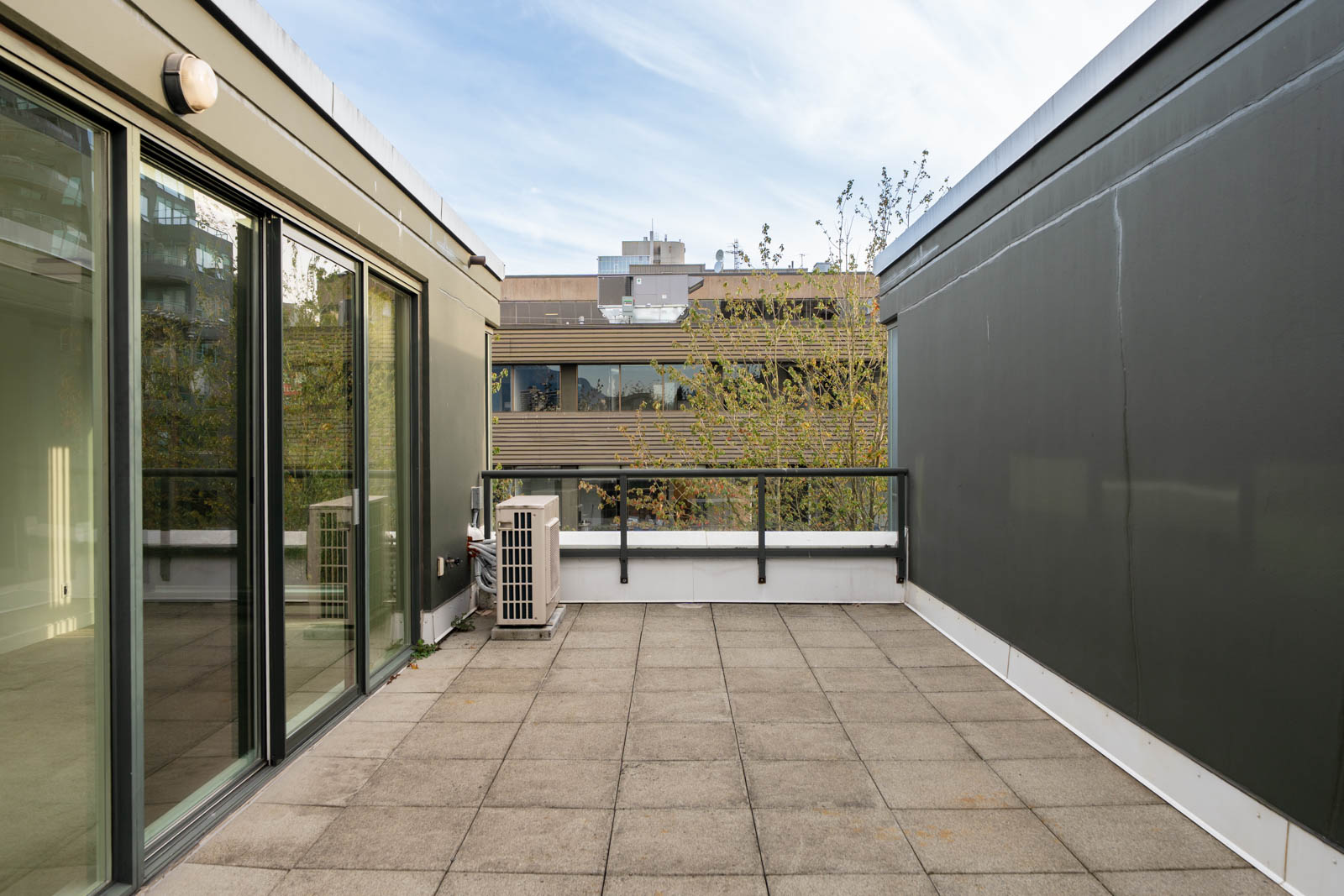 Outdoor terrace with tiled floors, glass sliding doors, air conditioning unit, and views of buildings and trees in Vancouver.