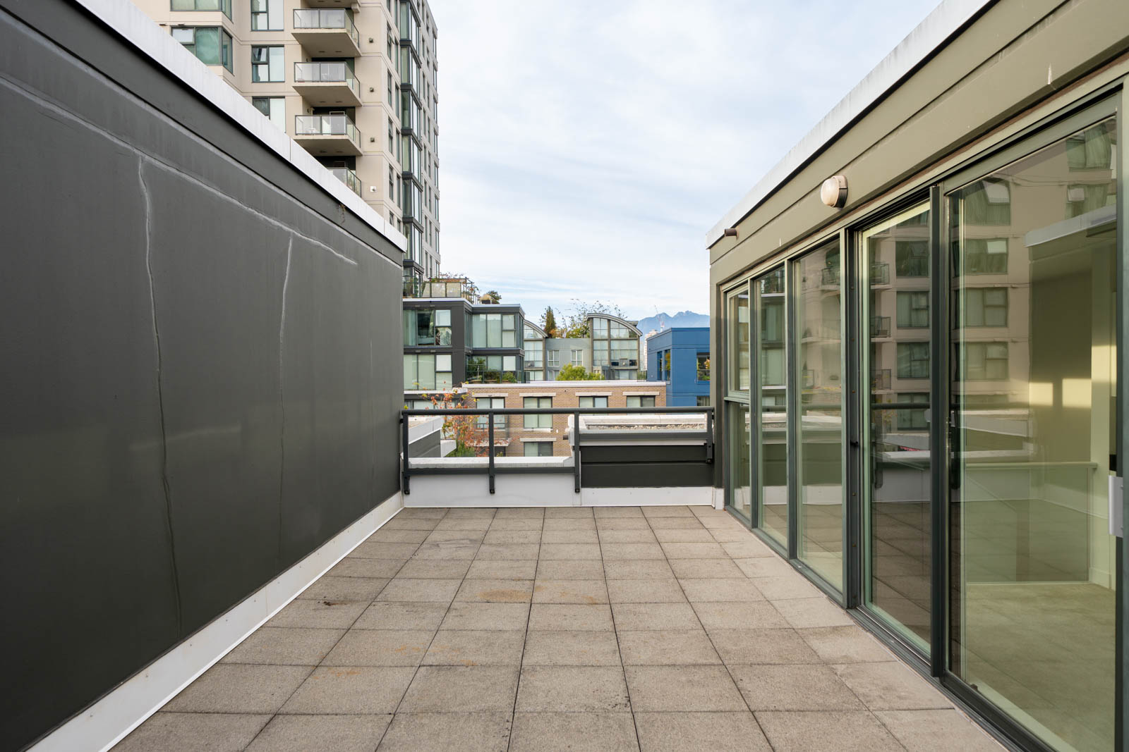 Terrace with tiled floor, glass sliding doors, and modern buildings nearby under a cloudy sky in a Vancouver West 6th Ave townhouse.