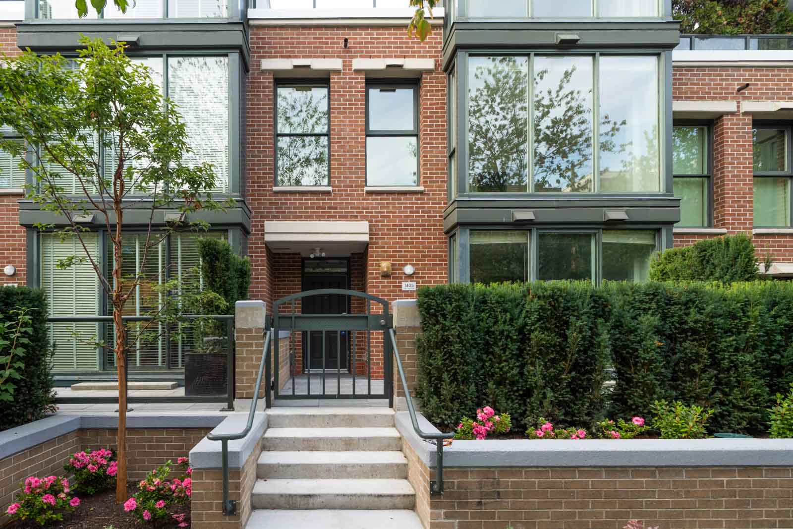 Modern townhouse at 1428 West 6th Ave, Vancouver, with large windows, gated entry, steps, green shrubs, and pink flowers in front.