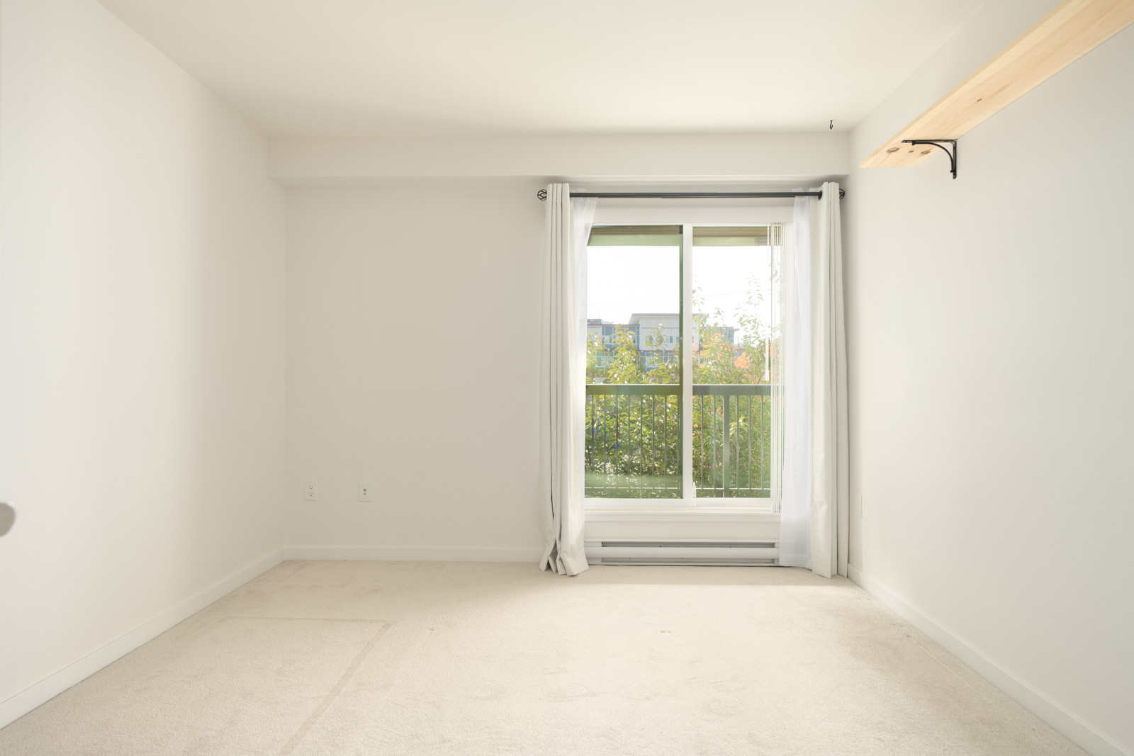 Carpeted room with white walls, a window with white curtains, wooden shelf on right, sunlight from greenery; Port Coquitlam near Wilson Ave.
