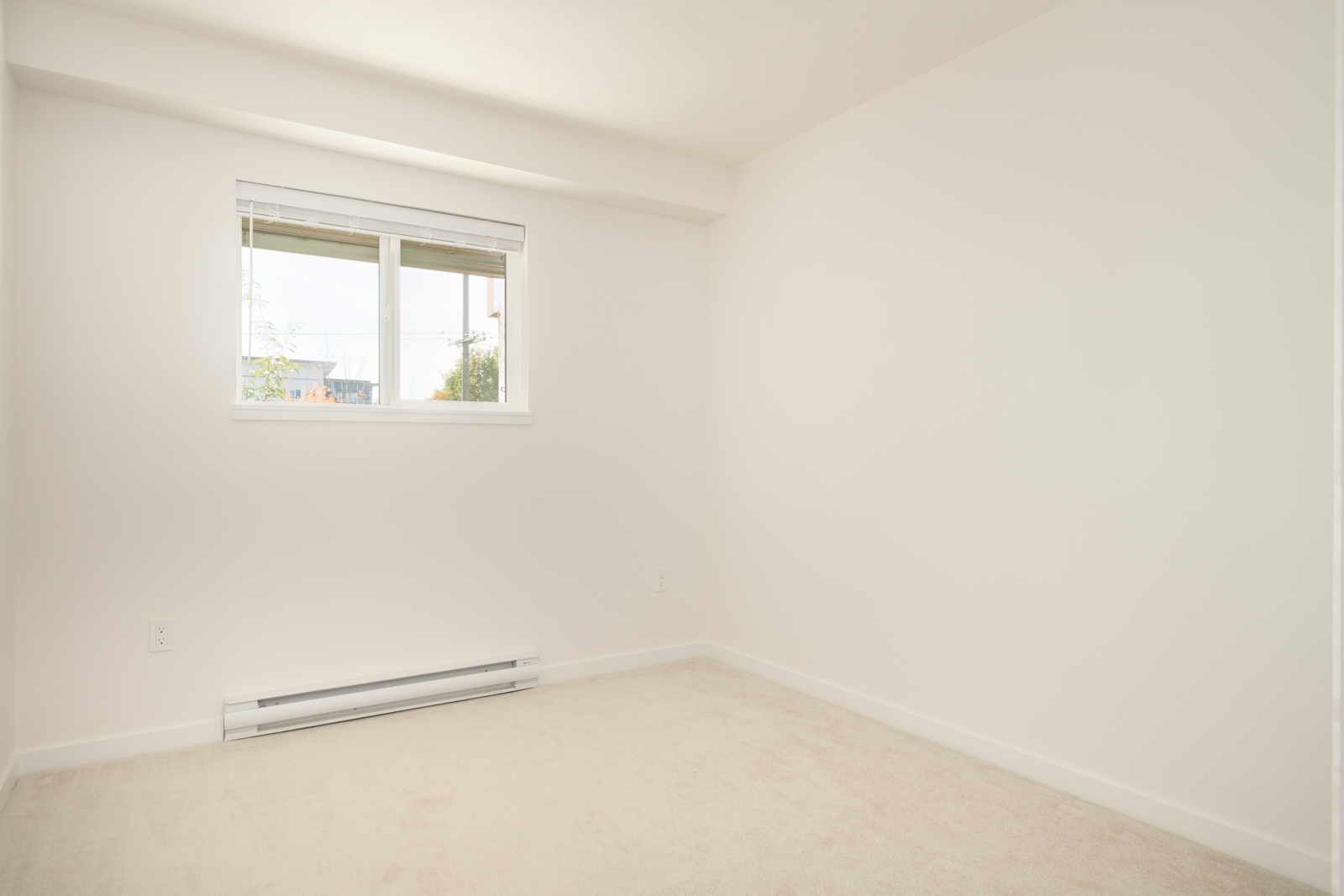 Empty 3rd-floor room with beige carpet, white walls, small window with blinds over Wilson Ave, baseboard heater below. Port Coquitlam.