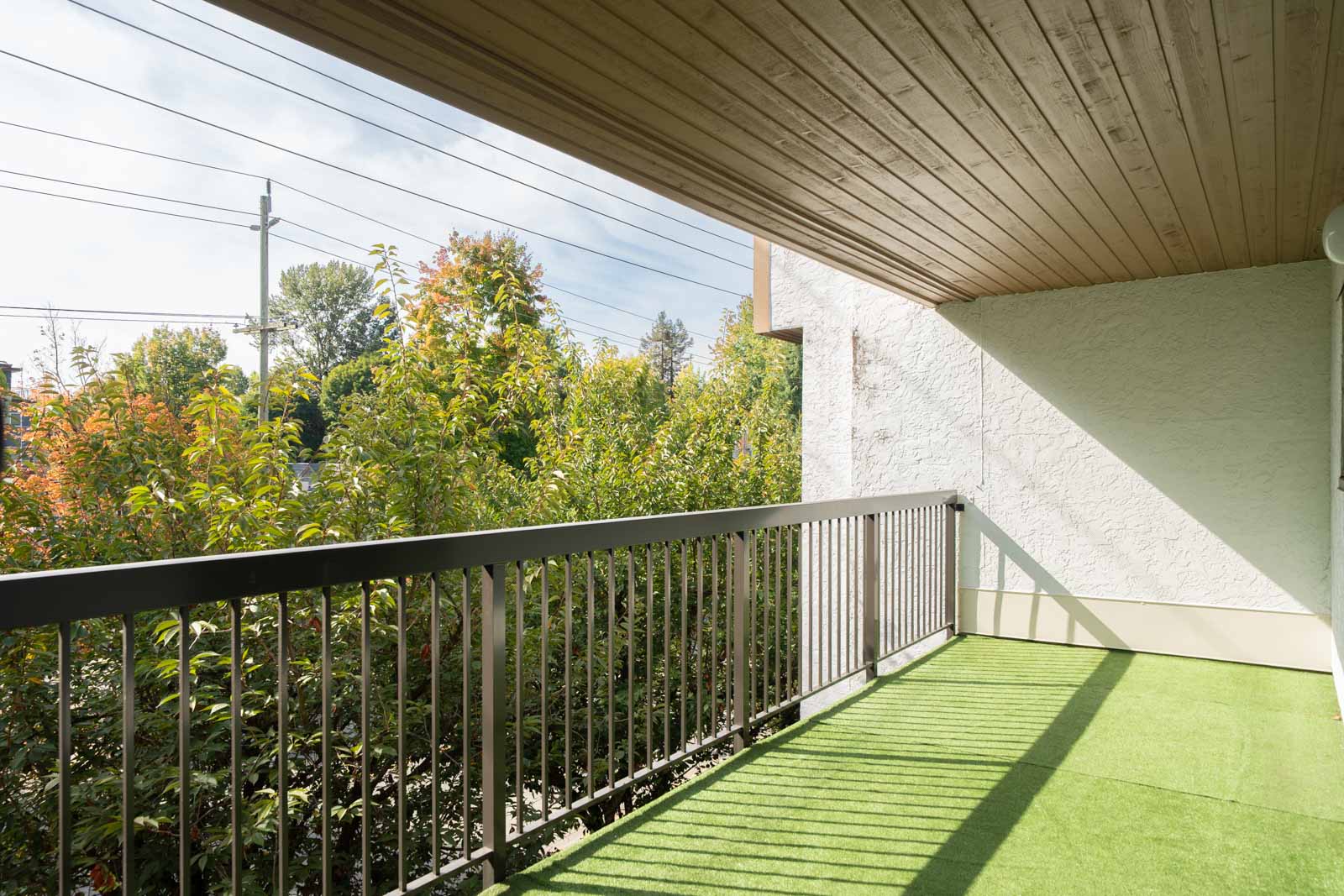 Covered 3rd floor balcony with green artificial grass, metal railing, and view of trees and power lines in Port Coquitlam.