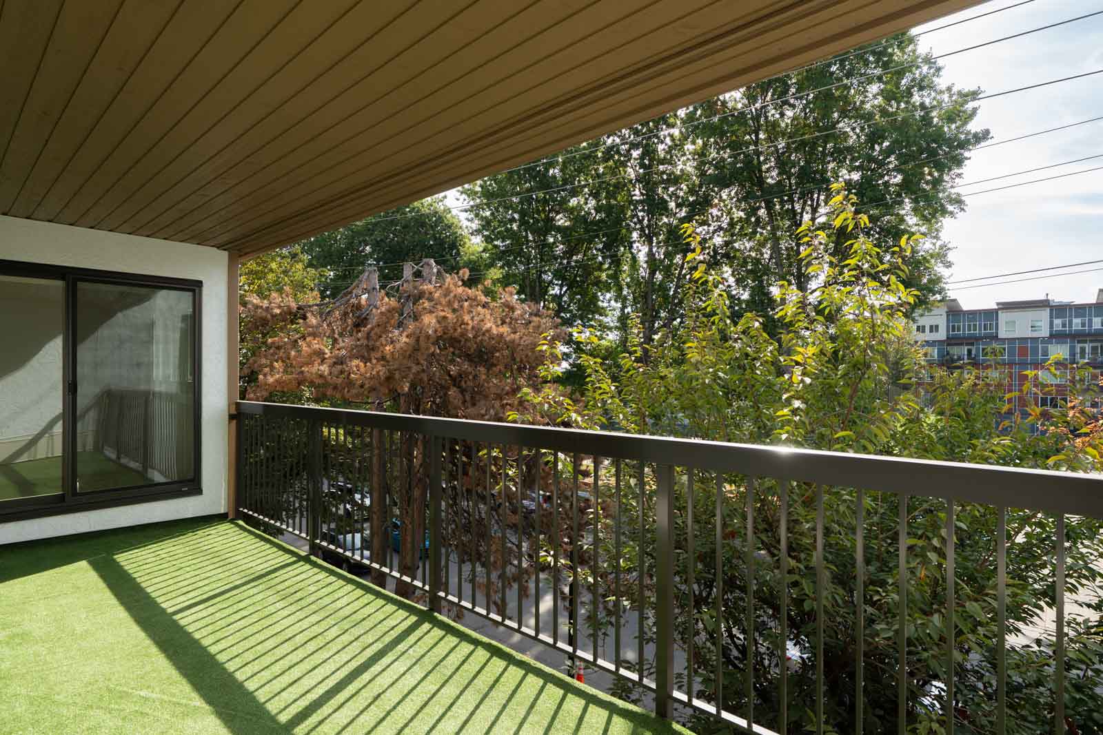 Covered balcony with artificial grass, metal railing, and large window overlooking trees and Wilson Ave in Port Coquitlam.
