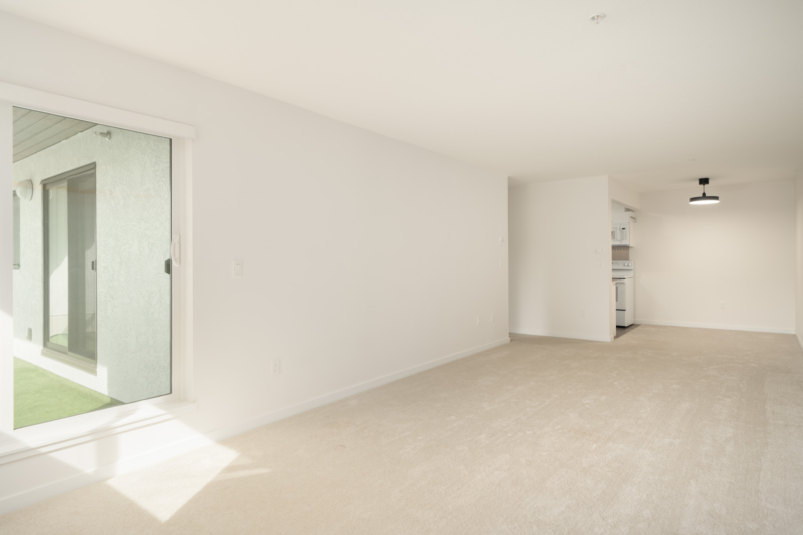Carpeted living room with white walls, large sliding glass door to patio, open to kitchen with white appliances in Port Coquitlam.