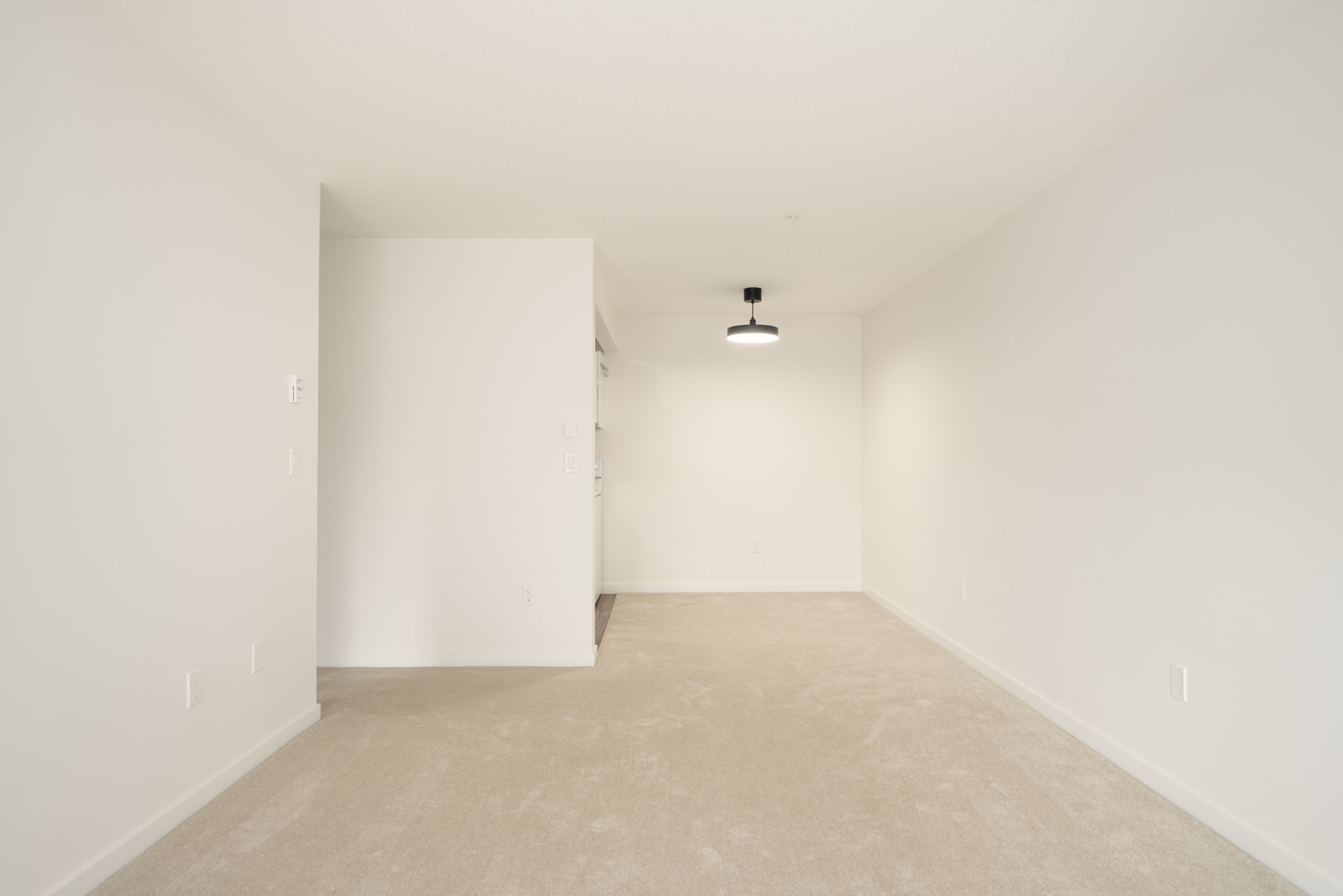 Empty room with beige carpet, white walls, and a modern ceiling light on the 3rd floor of a Wilson Ave building in Port Coquitlam; open doorway on left.