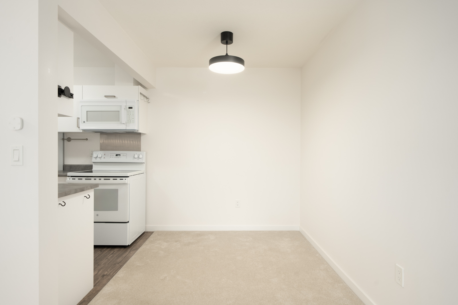 Small kitchen with white cabinets, stove, and microwave by an empty dining area with beige carpet and modern ceiling light in Port Coquitlam.