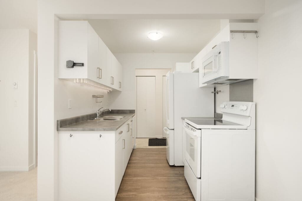 A galley kitchen at 2245 Wilson Ave, Port Coquitlam with white cabinets, grey counters, white appliances, and wood-look floors.