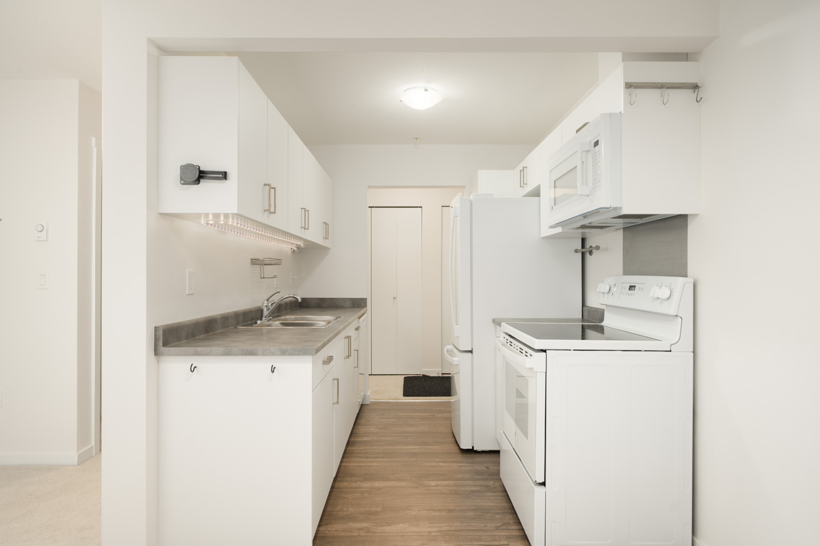 A galley kitchen at 2245 Wilson Ave, Port Coquitlam with white cabinets, grey counters, white appliances, and wood-look floors.