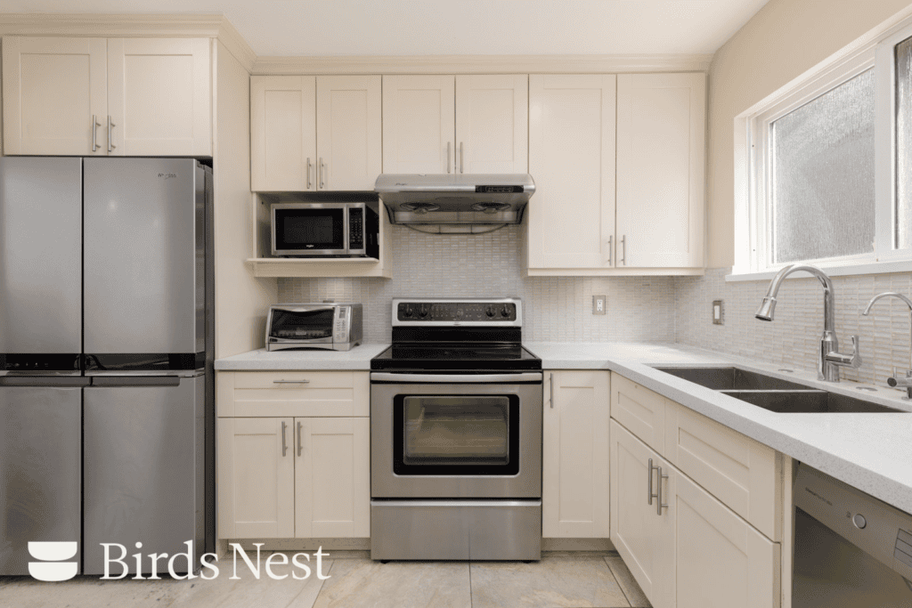 Modern kitchen near West 8th Ave, Vancouver, with white cabinets, stainless appliances, double sink, and “Birds Nest” logo in corner.