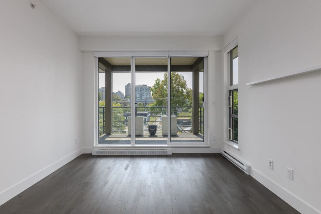 Empty room with dark wood floor, white walls, large windows, and glass door to balcony with seating and city view in Vancouver.