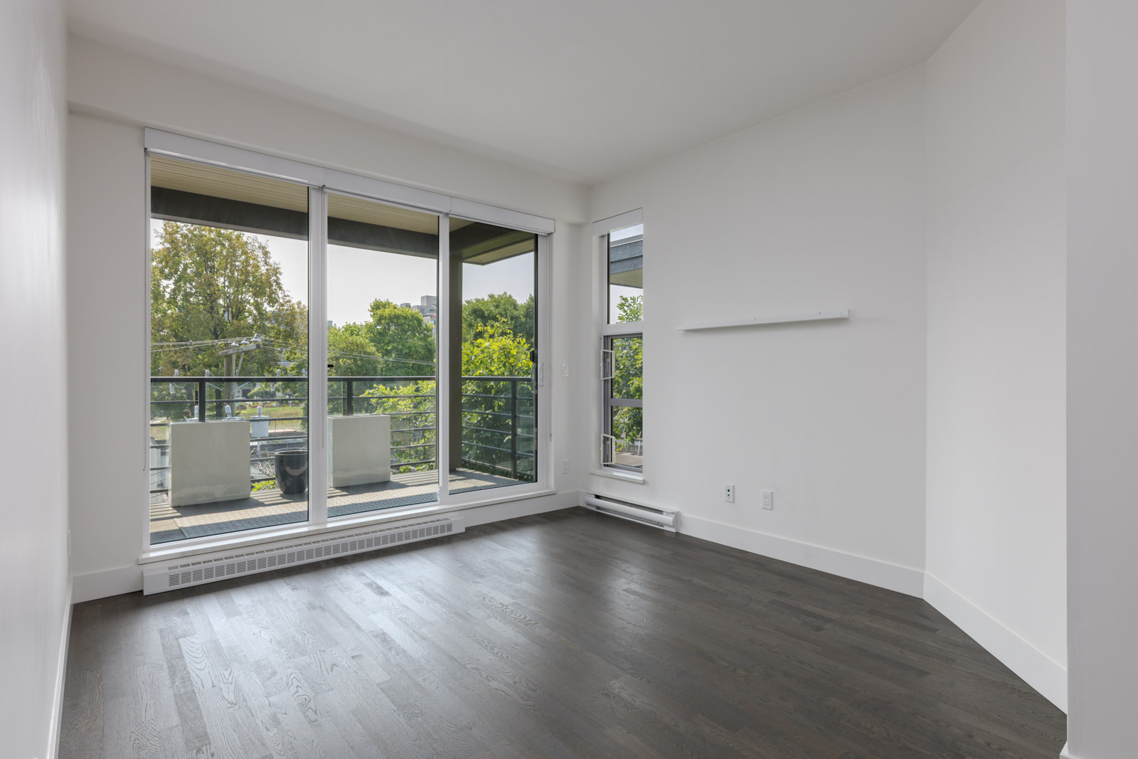 Empty room with dark wood floors, white walls, large windows, and glass door to balcony overlooking trees in Vancouver.