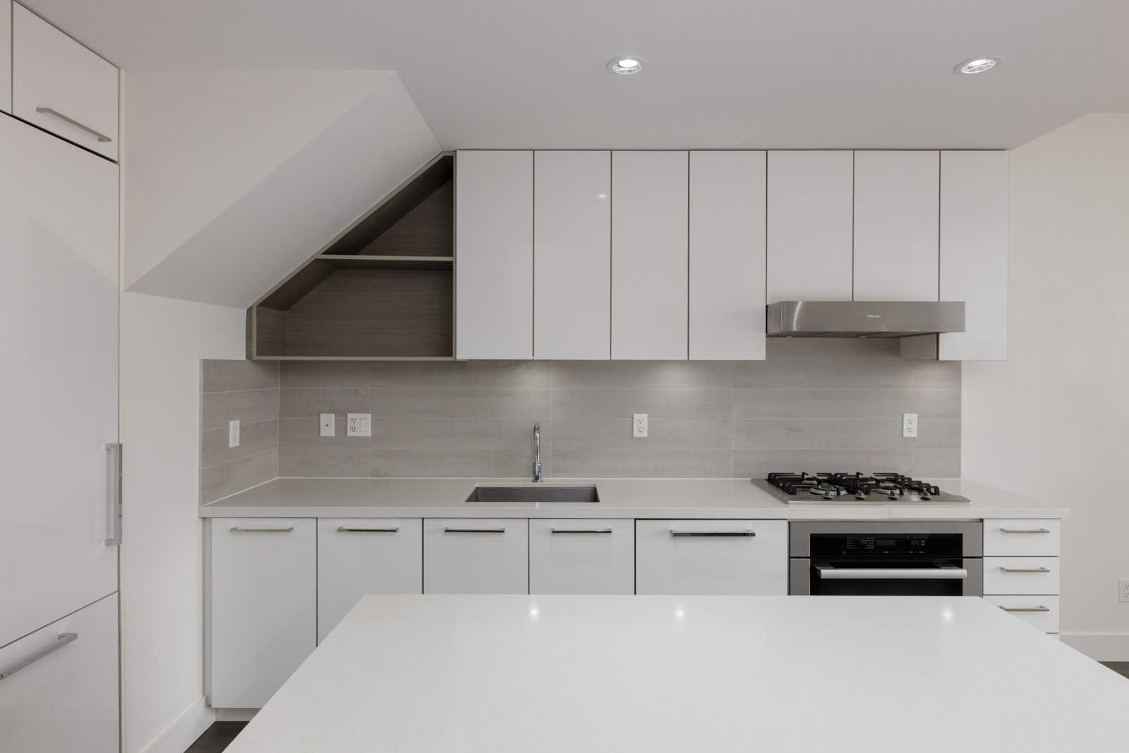 Modern kitchen with white cabinets, built-in stove, stainless hood, tiled backsplash, island, and open corner shelves in Vancouver.