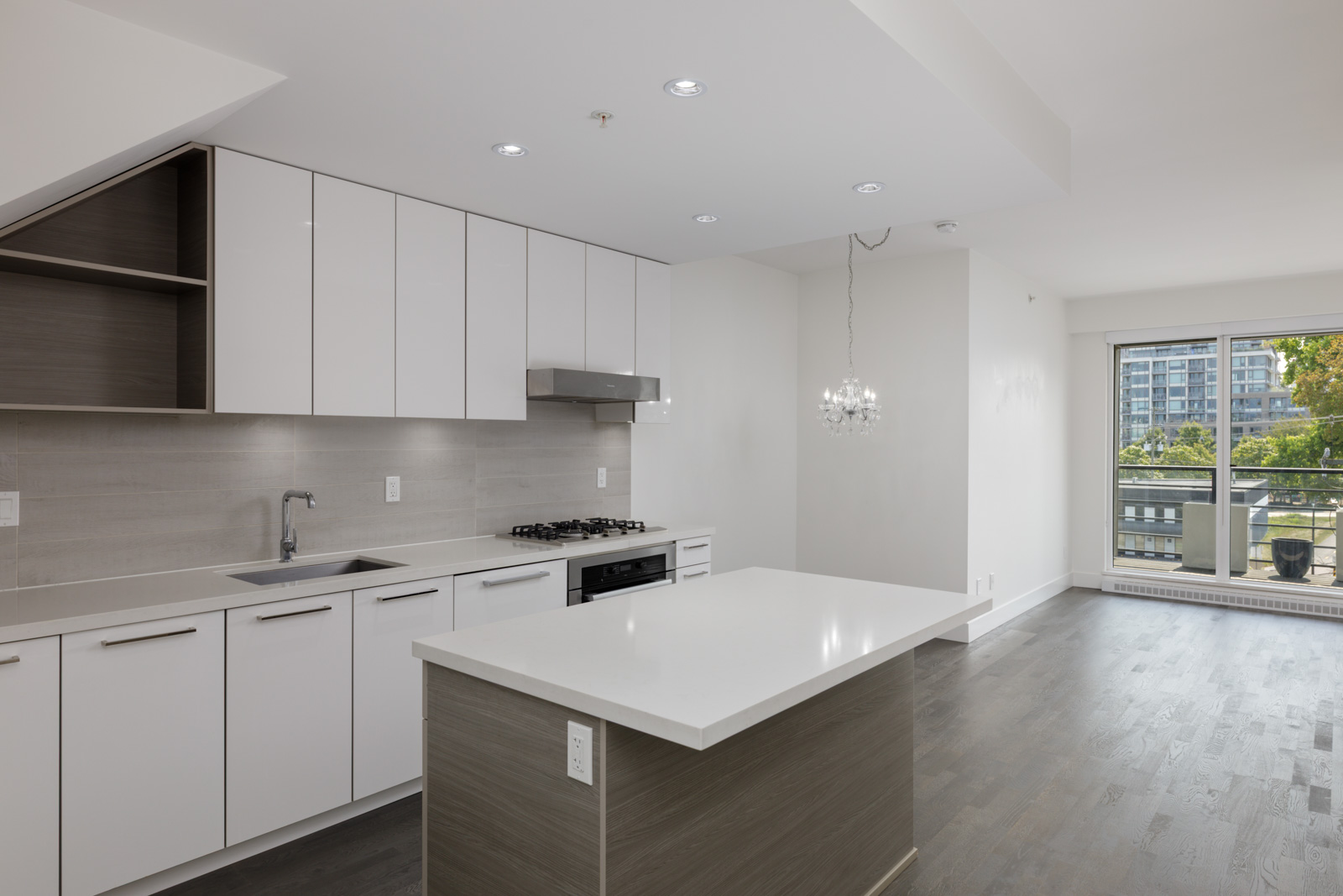 Modern kitchen with white cabinets, center island, built-in stove, open living space, large window and balcony view in Vancouver.