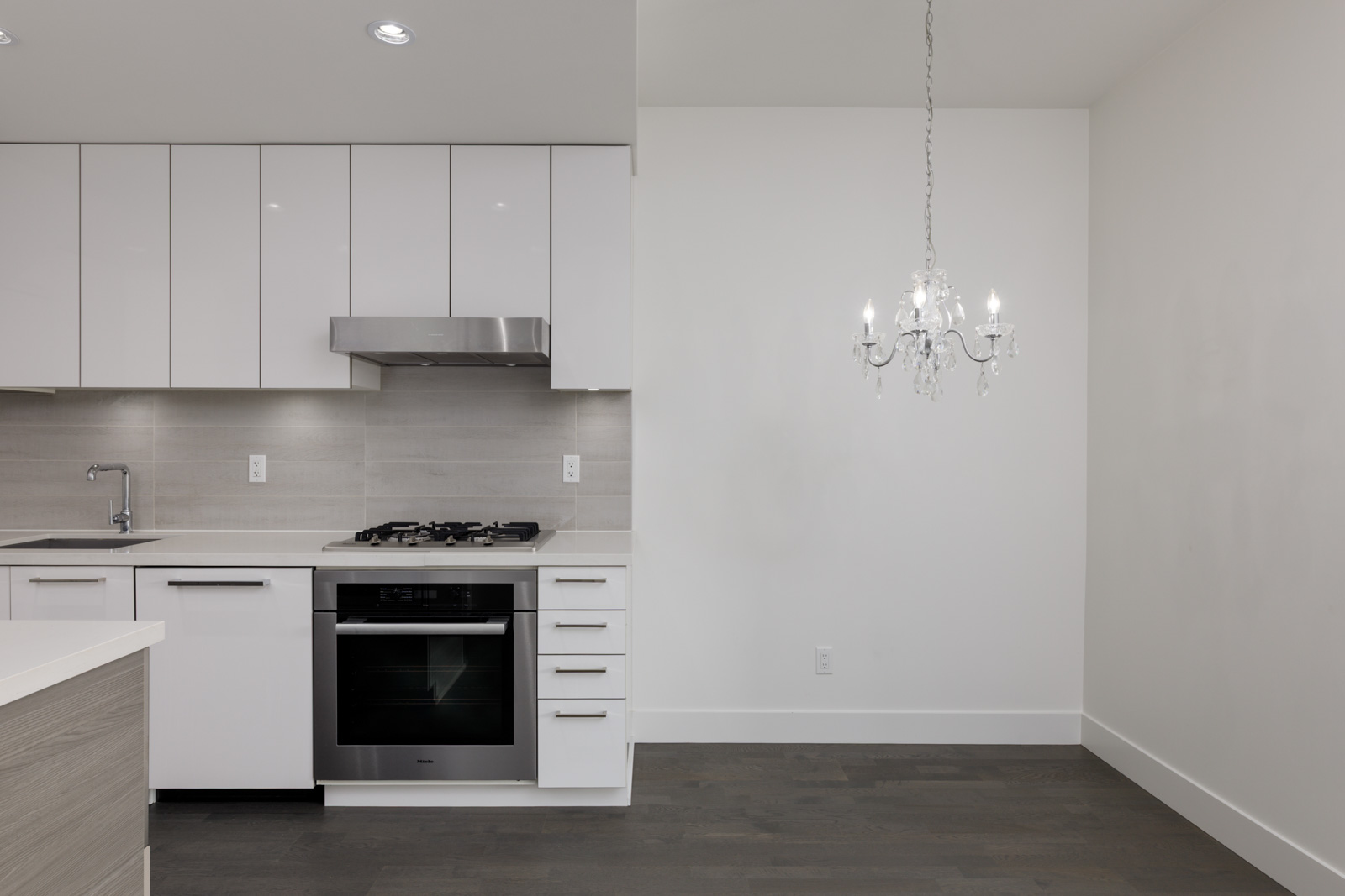 Modern kitchen at 1628 W 4th Ave, Vancouver, with white cabinets, stainless appliances, gray backsplash, and chandelier over dark wood floor.