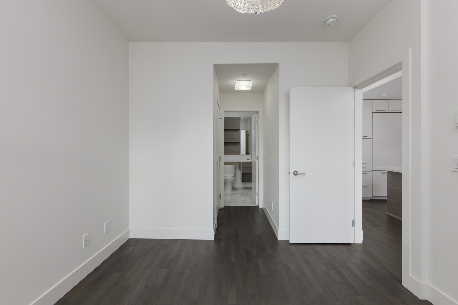 Modern, empty room with white walls, dark wood floor, open bathroom door, and kitchen view; 4th Floor, 1628 W 4th Ave, Vancouver.