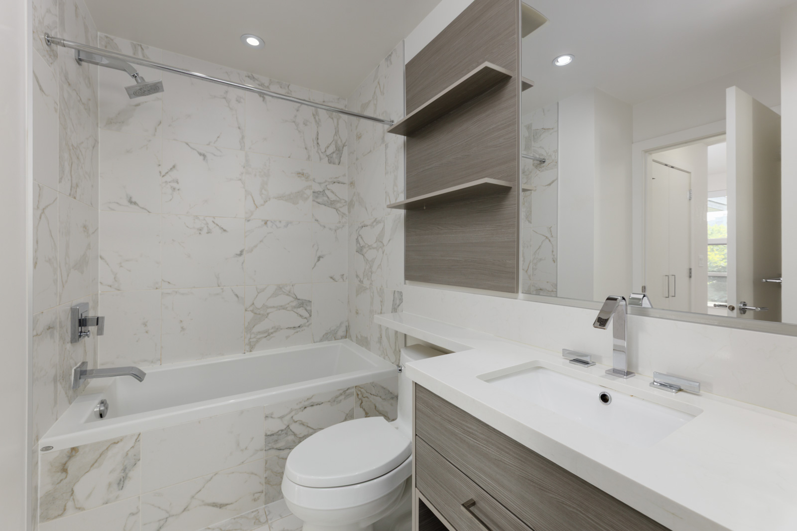 Modern bathroom on 4th Floor at 1628 W 4th Ave, Vancouver; has a tub-shower, white marble tiles, wood cabinetry, and large mirror.