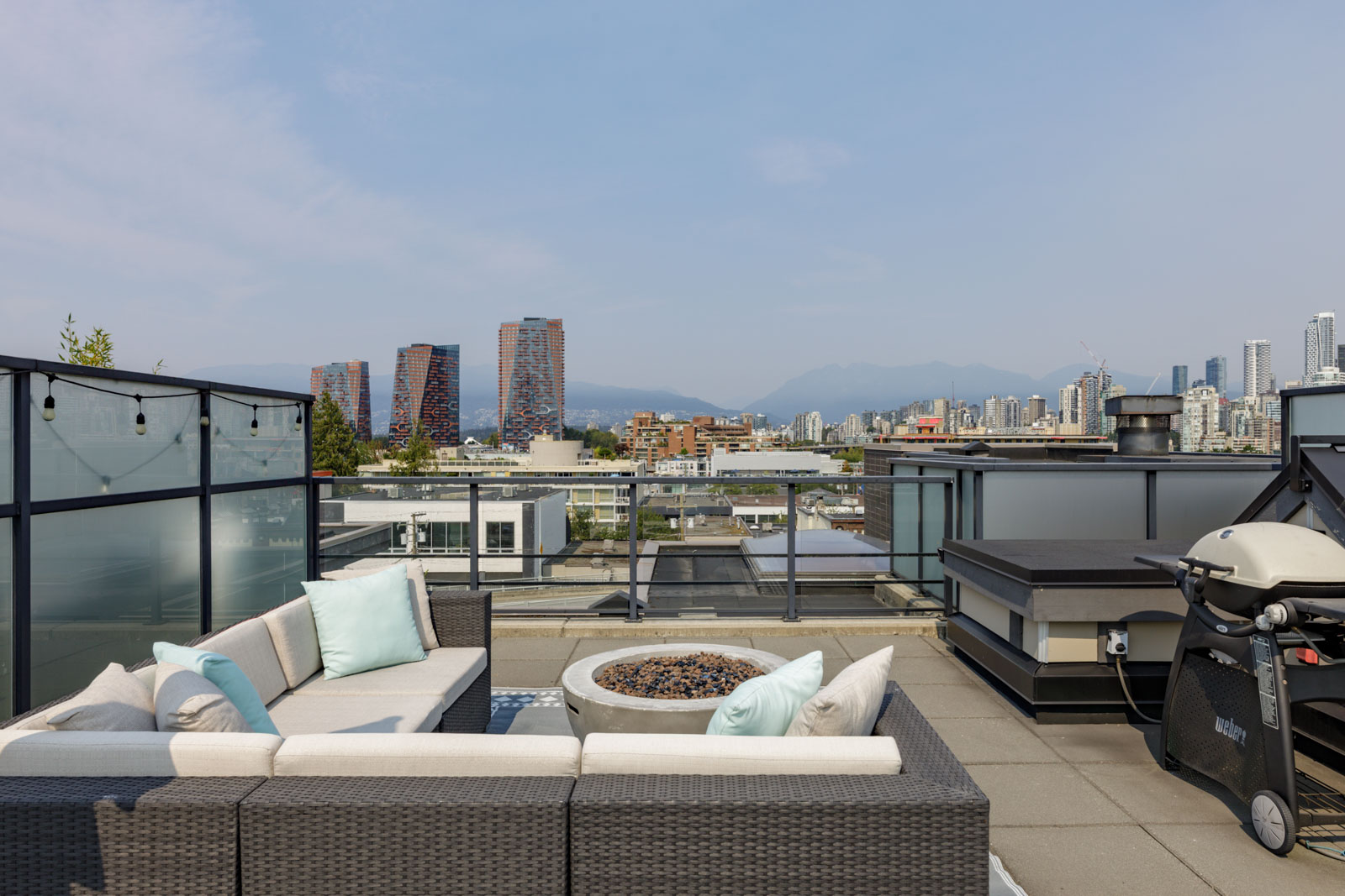 Rooftop patio on 4th floor, 1628 W 4th Ave, Vancouver, with wicker seating, fire pit, grill, and city skyline plus mountains.