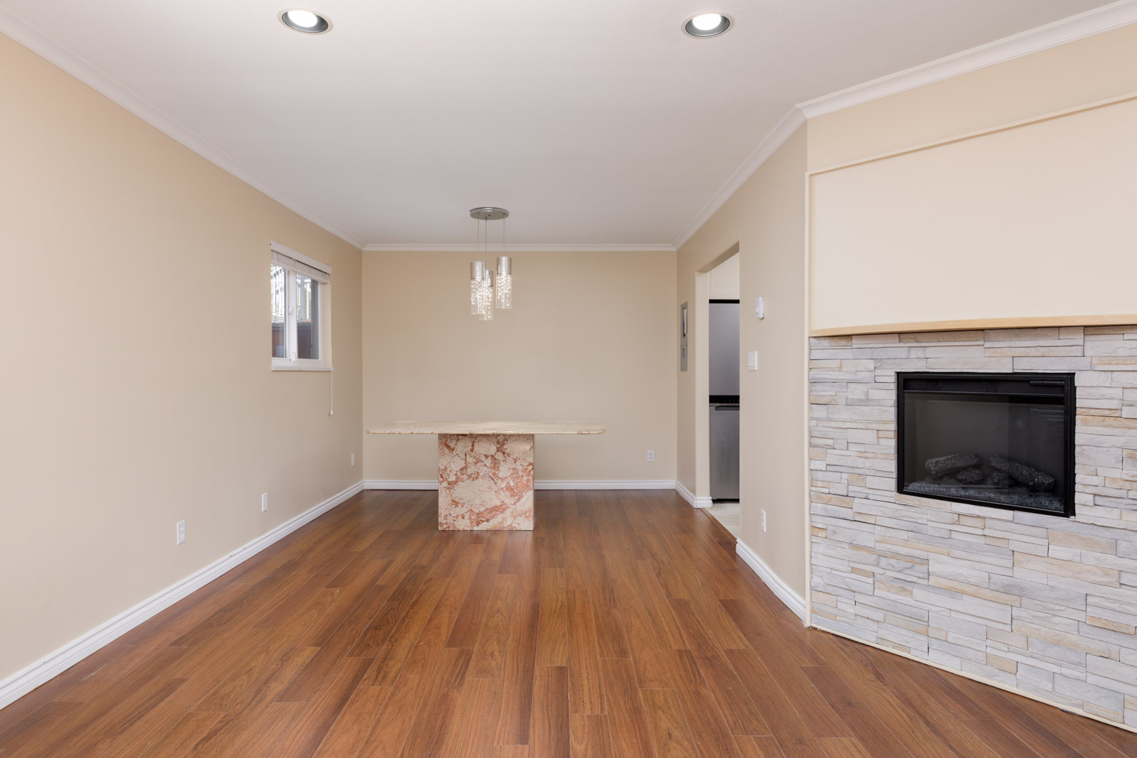 Empty room with wood floors, beige walls, stone fireplace right, small window left, marble-base table, chandelier; Vancouver.