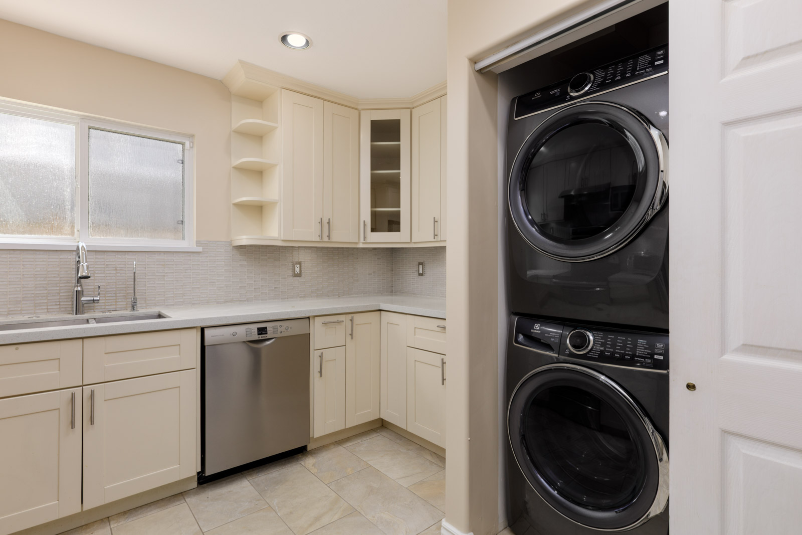 Modern laundry room with cream cabinets, stainless steel dishwasher, sink under window, stacked washer-dryer by sliding door in Vancouver.