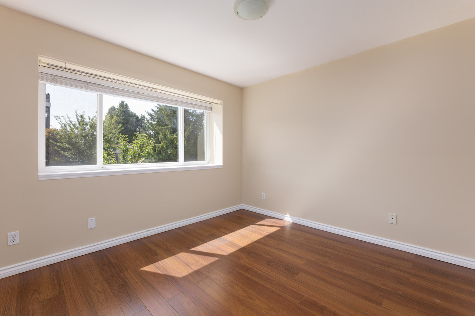 Empty room with beige walls, wood floors, big window with white blinds, sun casting shadows—Musqueamview St near W 8th Ave, Vancouver.