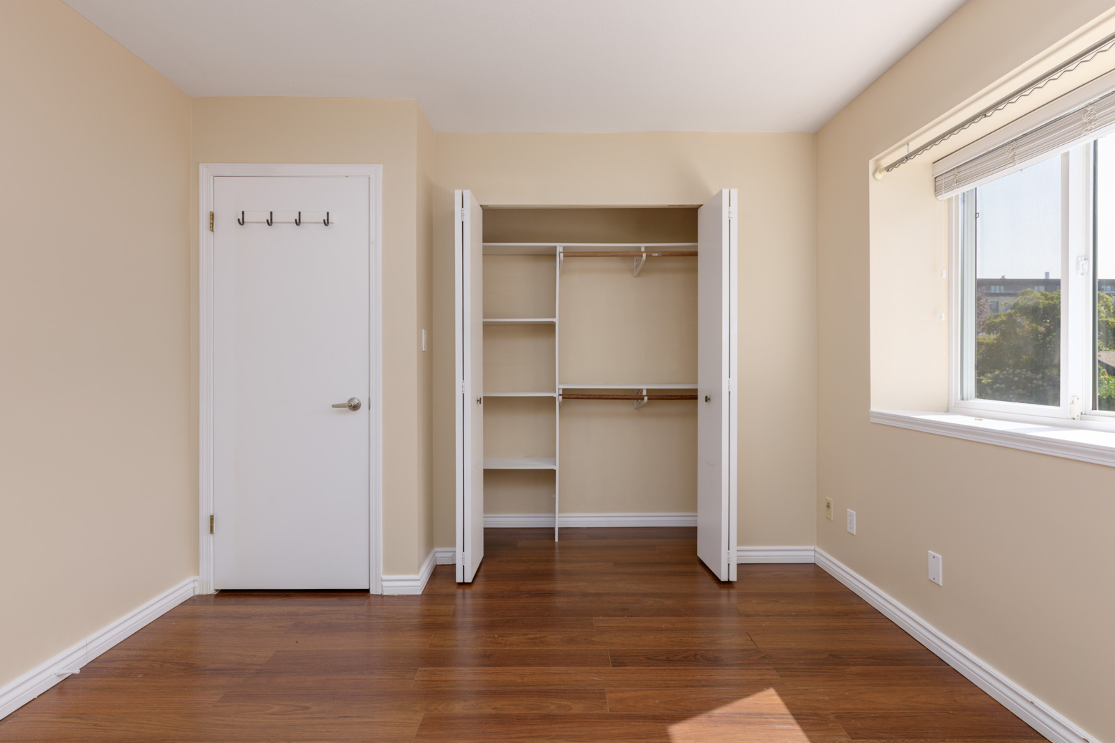 Empty room with beige walls, open closet, white door with hooks, wood floor, and large window in a Musqueamview St building, Vancouver.