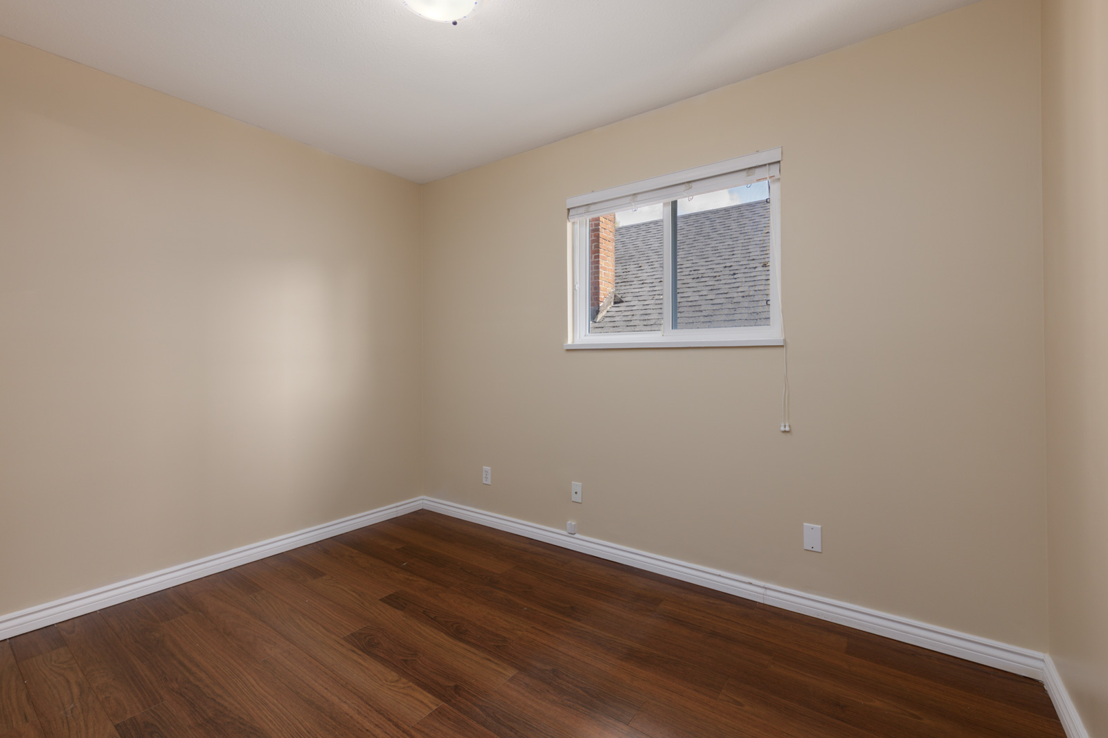 Empty room with beige walls, white trim, hardwood floor, ceiling light, and window overlooking a roof near West 8th Ave, Vancouver.