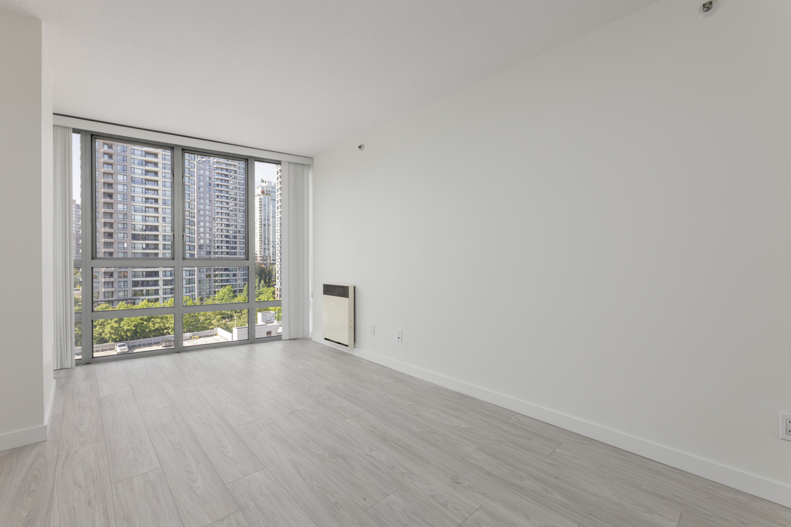 Bright, empty room with light wood floors, white walls, and large windows at 950 Cambie St, Vancouver overlooking city and trees.